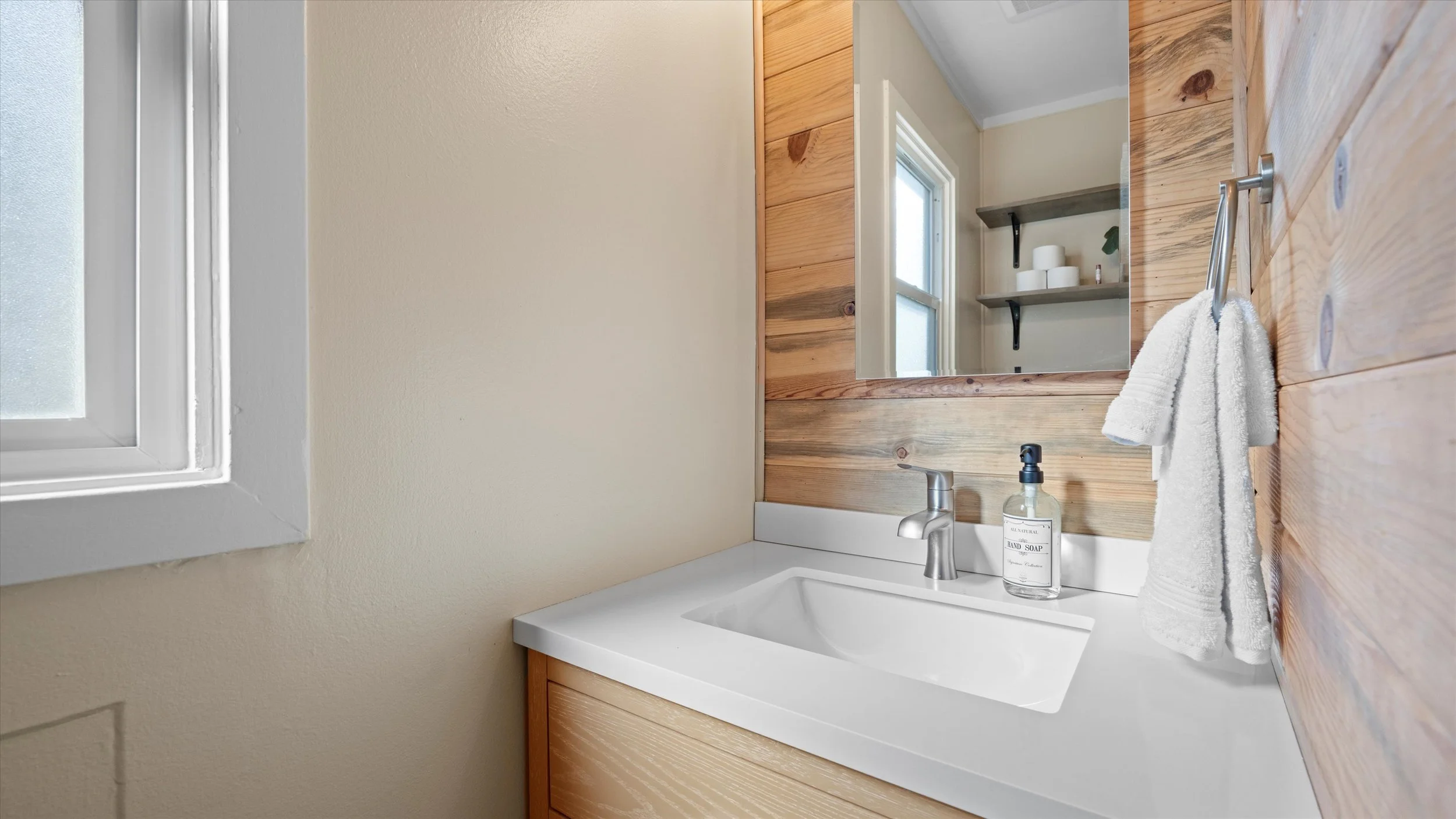 Bathroom sink area with a white countertop, a silver faucet, hand soap in a bottle, a towel hanging on a hook, a wooden mirror frame, a window, and wooden shelves with rolled towels.