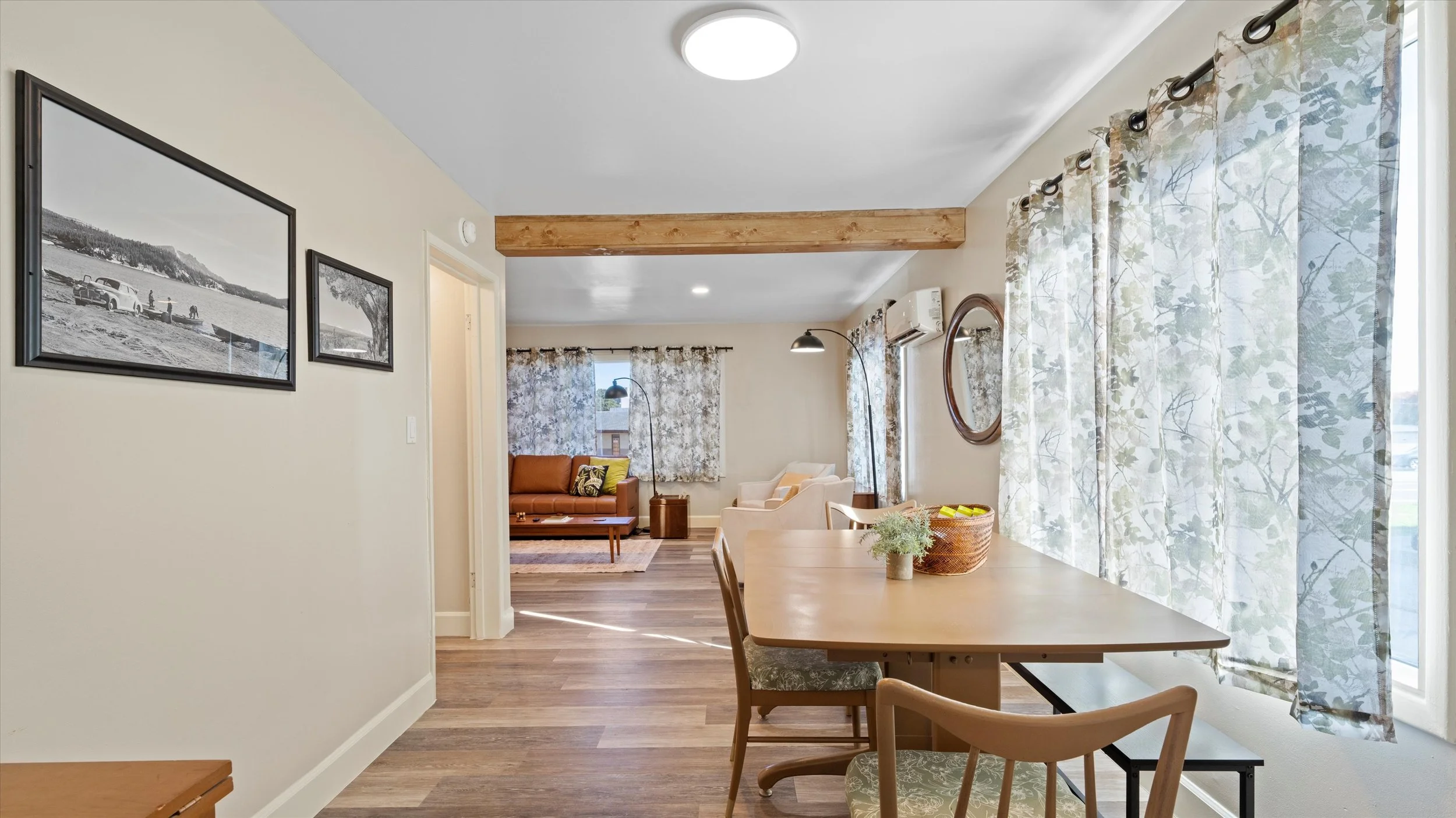 A view from the dining area into the living room of a home, with a wooden dining table, chairs, and decorative curtains on the windows.