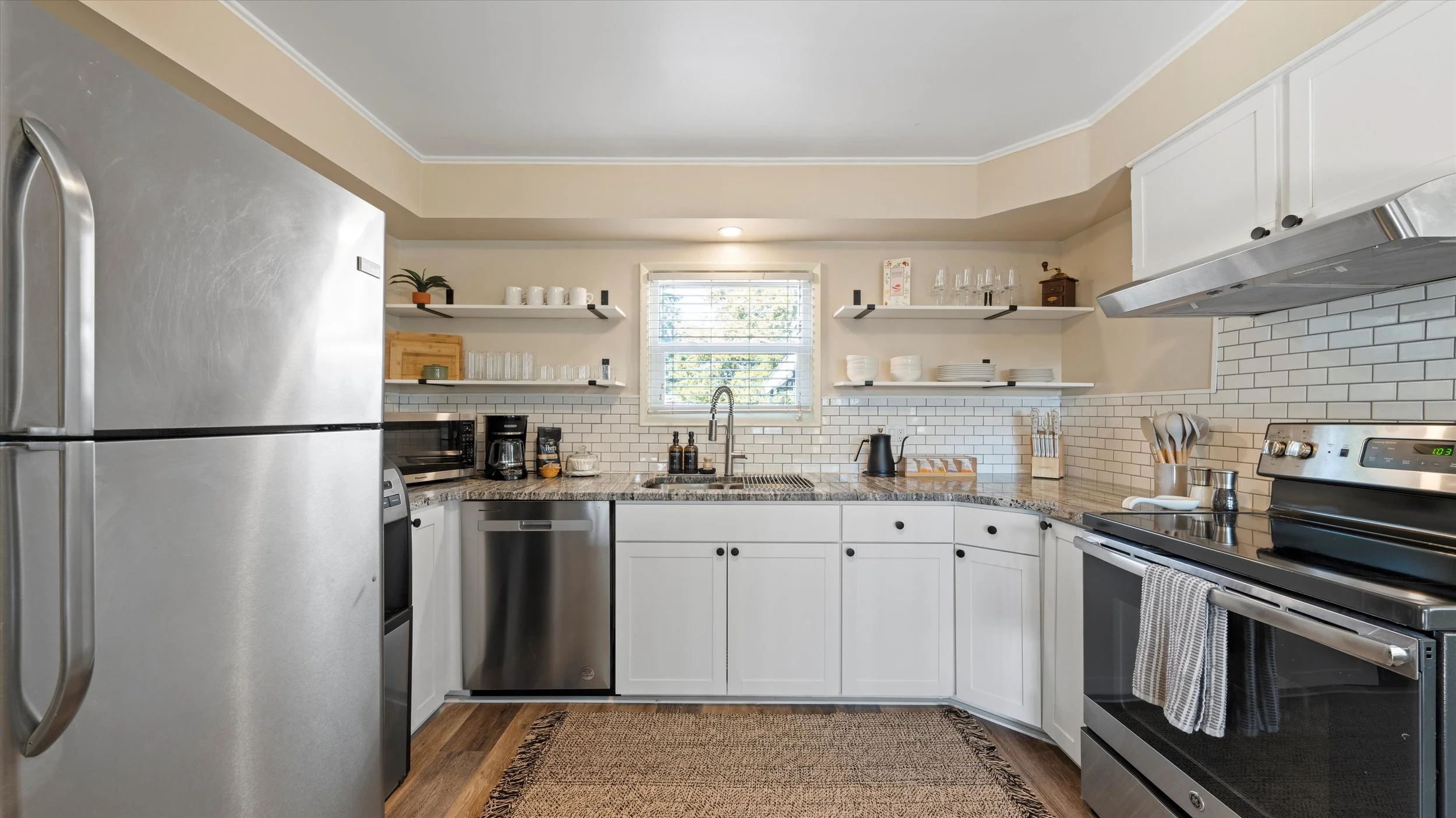 A modern kitchen with white cabinets, stainless steel appliances, open shelves with dishes and glasses, a granite countertop, and a window above the sink.