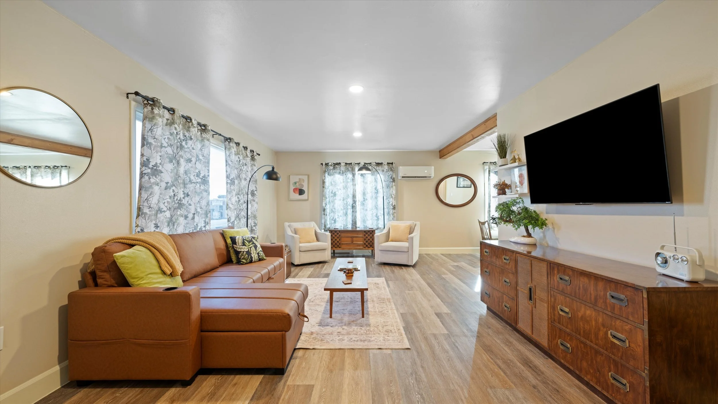 Spacious living room with beige walls, large windows with floral curtains, a brown leather sofa with yellow and black pillows, two white armchairs with yellow pillows, a wooden coffee table, a large wall-mounted TV, a wooden sideboard with a bonsai t
