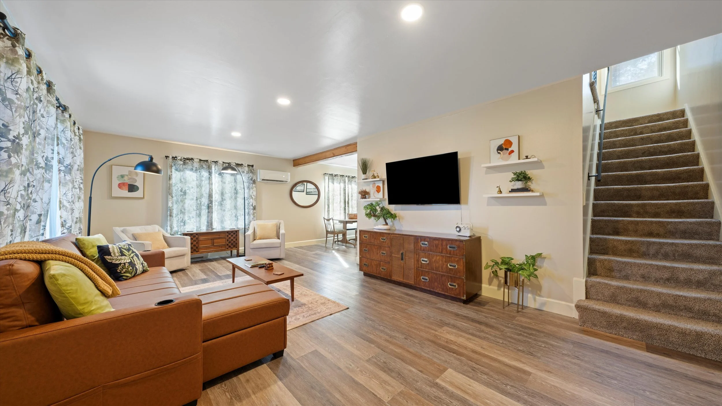 Living room with brown leather sofa, beige armchair, wooden coffee table, flat-screen TV, shelves with decor, staircase with carpet, hardwood flooring, and windows with patterned curtains.