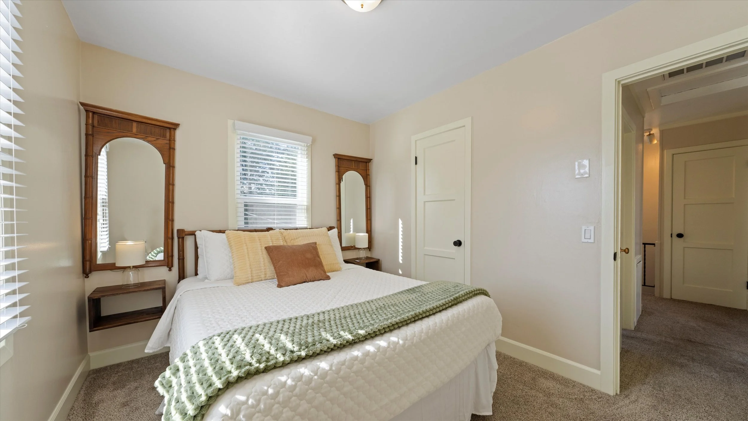 A cozy bedroom with a neatly made bed featuring cream-colored and brown pillows, a green textured blanket, and a wooden headboard with mirrors on each side. There are two small nightstands with lamps, a window with blinds, and cream-colored walls wit