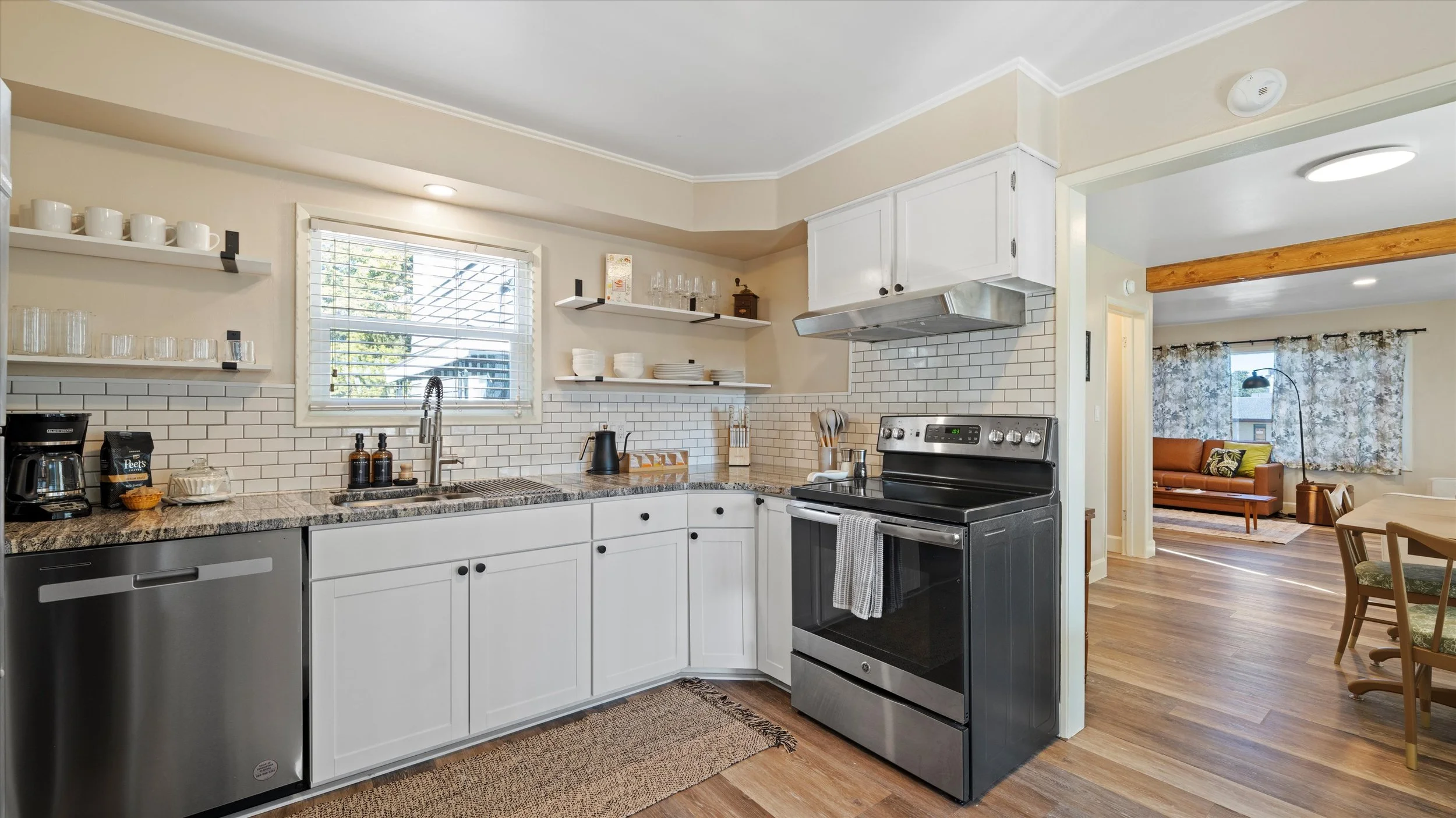 A kitchen with white cabinets, open shelves with dishes and glassware, a granite countertop with small appliances and utensils, a black stove, stainless steel dishwasher, and a window with blinds. Adjacent to the kitchen is a living area with a brown
