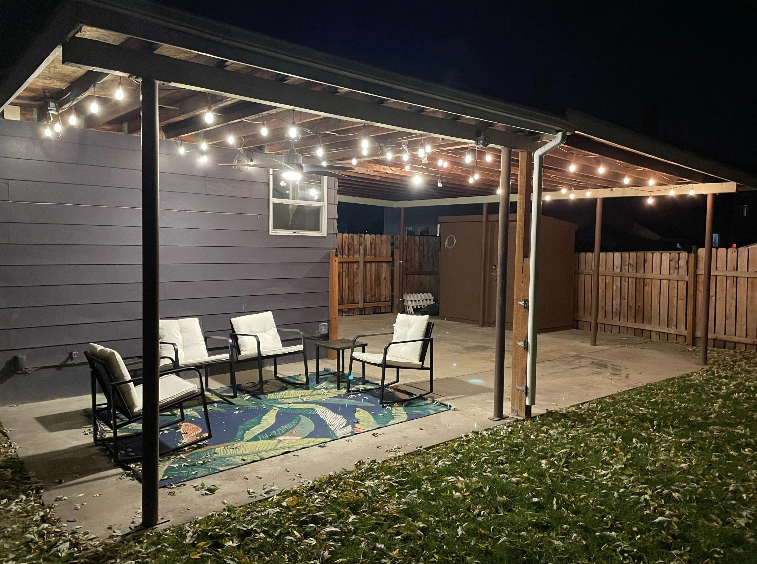 A backyard patio at night with string lights hanging from a wooden pergola, outdoor chairs, a patterned outdoor rug, a fence, and a shed.