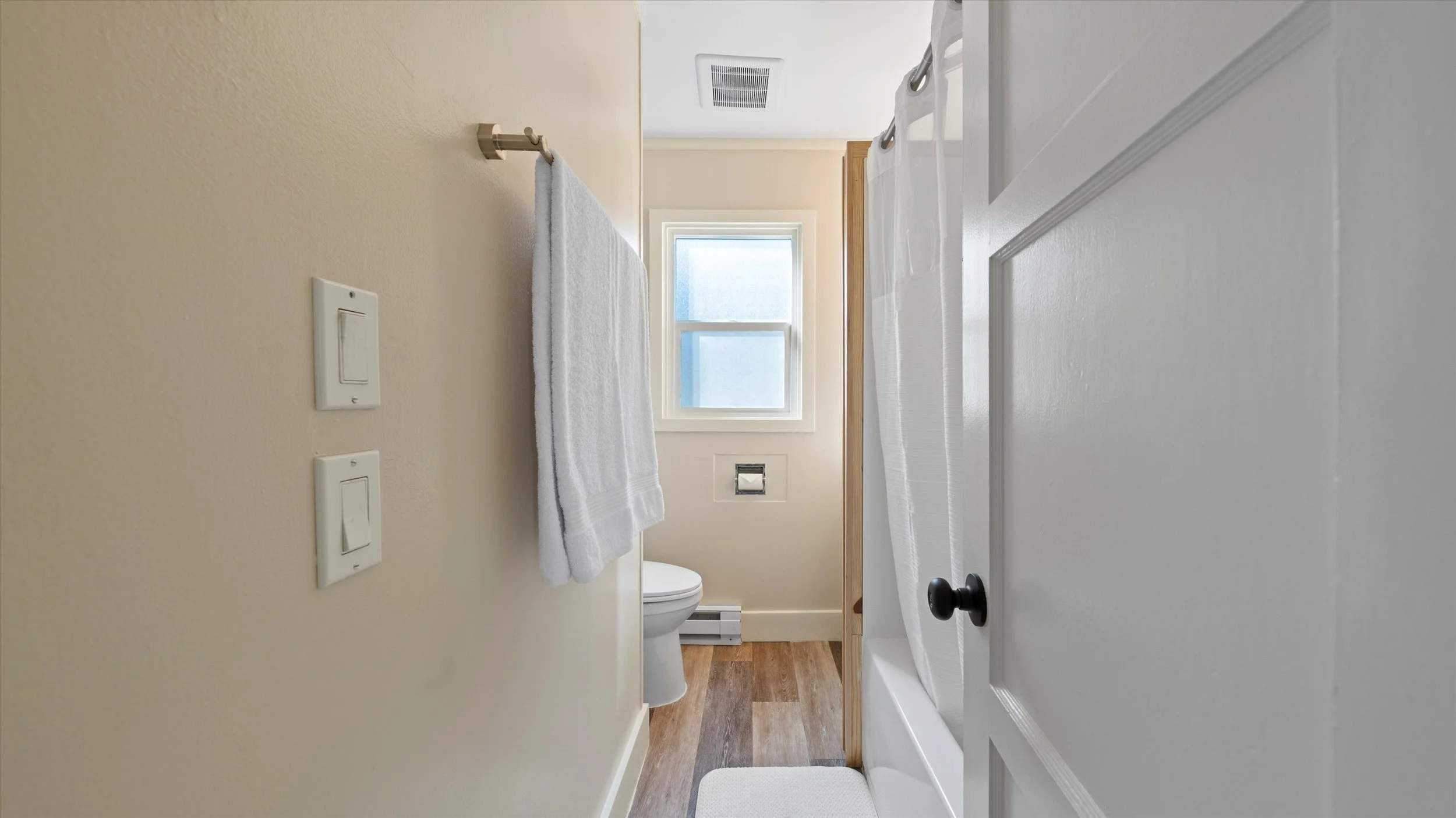 A small bathroom with a toilet, a window with frosted glass, a towel rack with a white towel, and a shower curtain. The walls are painted beige, and the floor is wood. The door is partly open.
