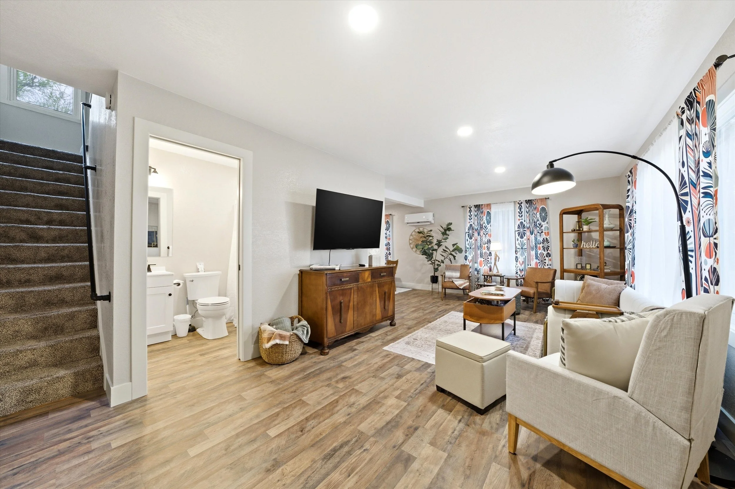 Living room with white walls and ceiling, a gray sofa, wooden furniture, colorful curtains, a flat-screen TV, and a staircase to the left. There is also a visible bathroom with a toilet and sink.