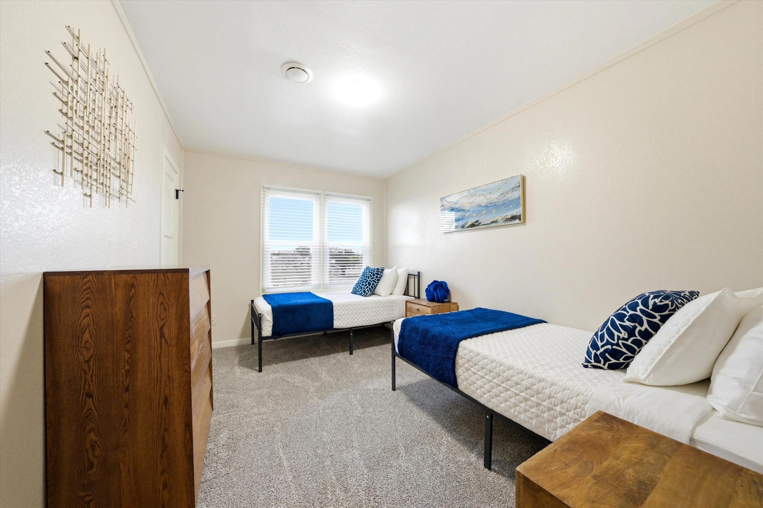 A bedroom with two twin beds covered in white bedding and blue throws, each with matching blue and white pillows. A small wooden nightstand with decorative blue objects separates the beds. There is a window with white blinds, a framed abstract sea painting on the wall, a textured metal wall art piece, a wooden dresser, and a ceiling light fixture.