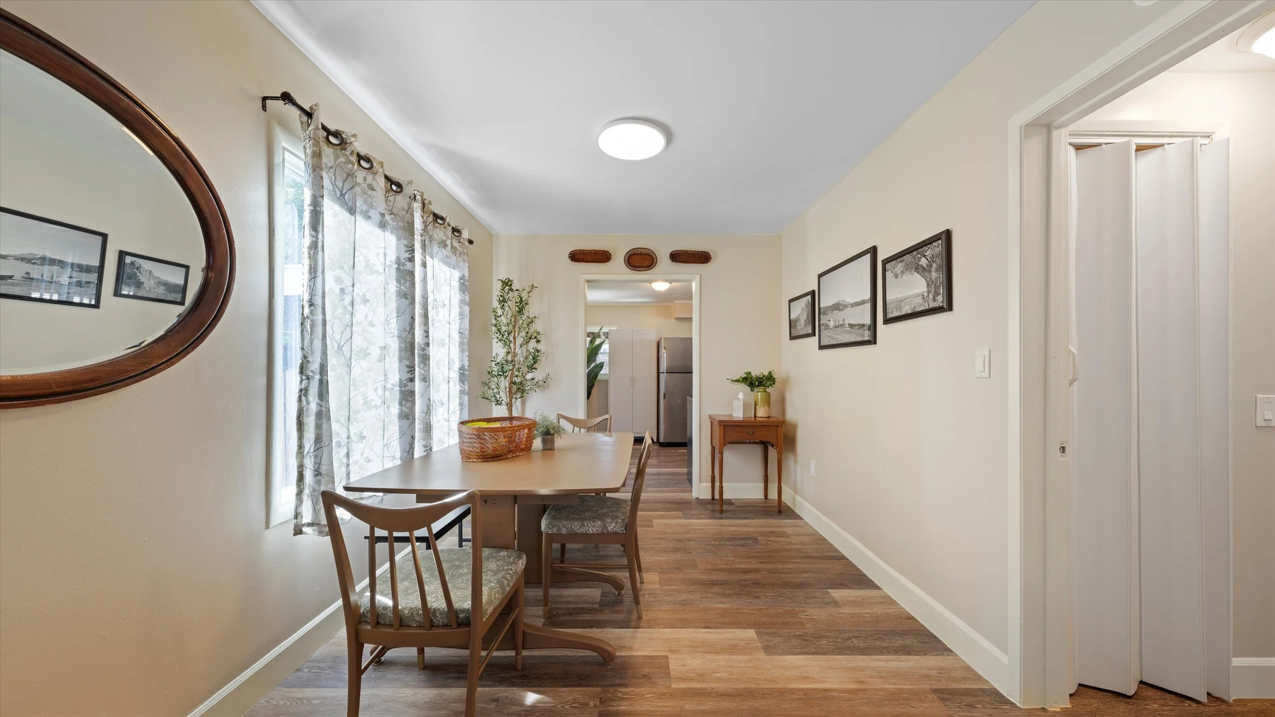 Dining area with wooden table and chairs, wall art, window with floral curtains, potted plant, additional furniture and kitchen in the background.
