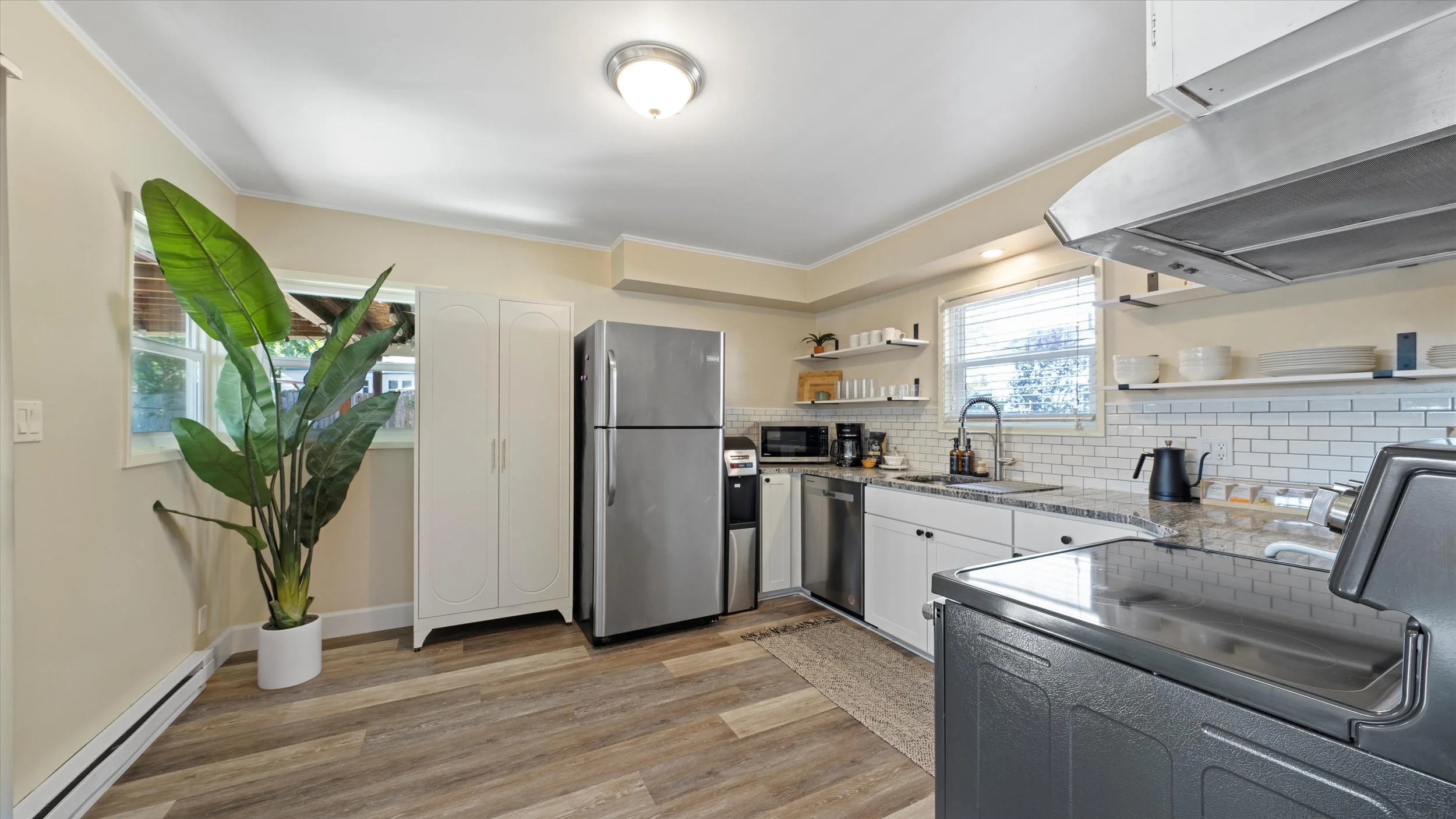 A modern kitchen with white cabinets, stainless steel refrigerator, granite countertops, open shelving, small appliances, and a tall potted plant near two small windows.