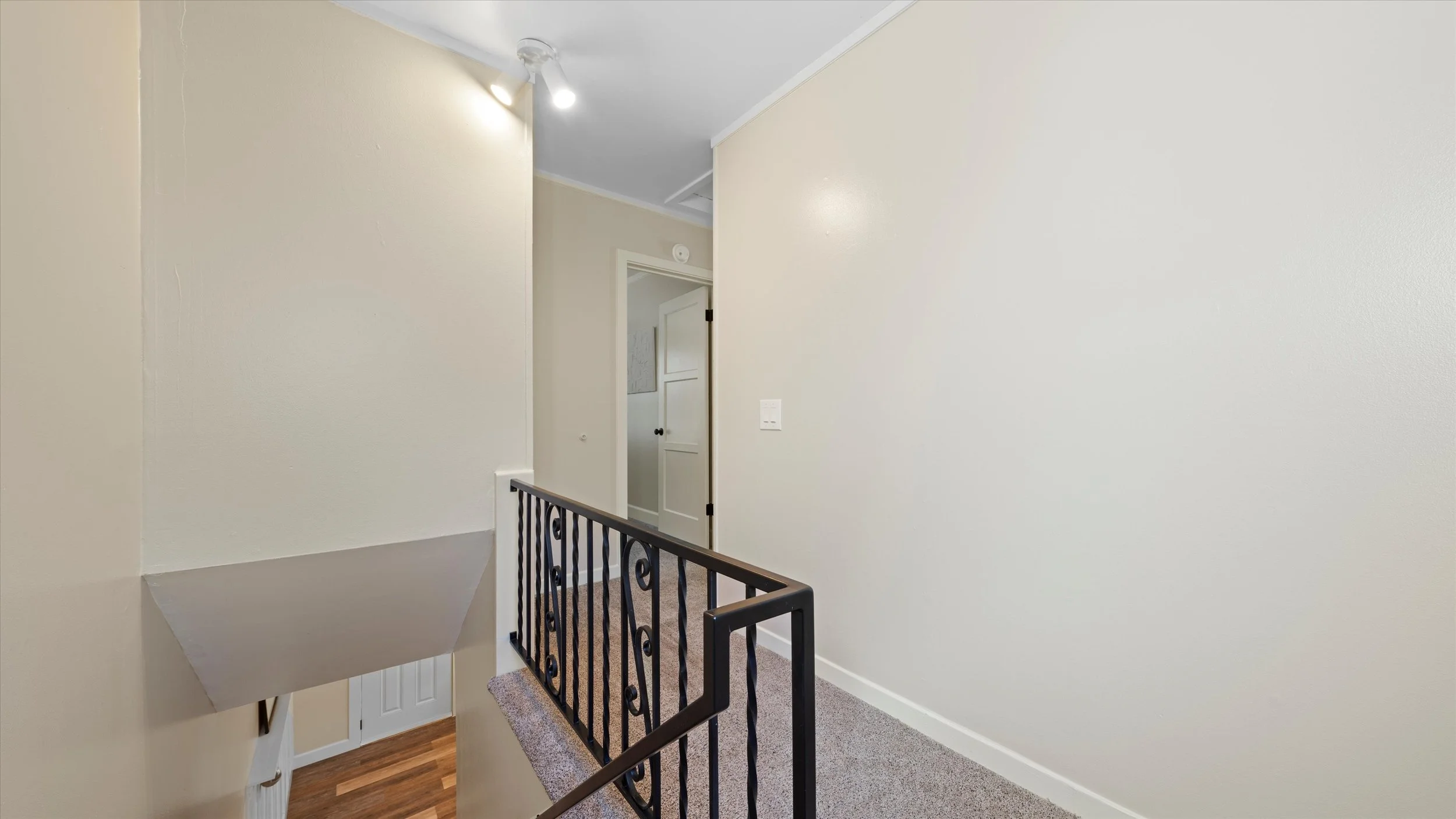 Interior view of a second-floor hallway with cream-colored walls, a small section of black wrought iron railing, a closed white door, and a ceiling light.