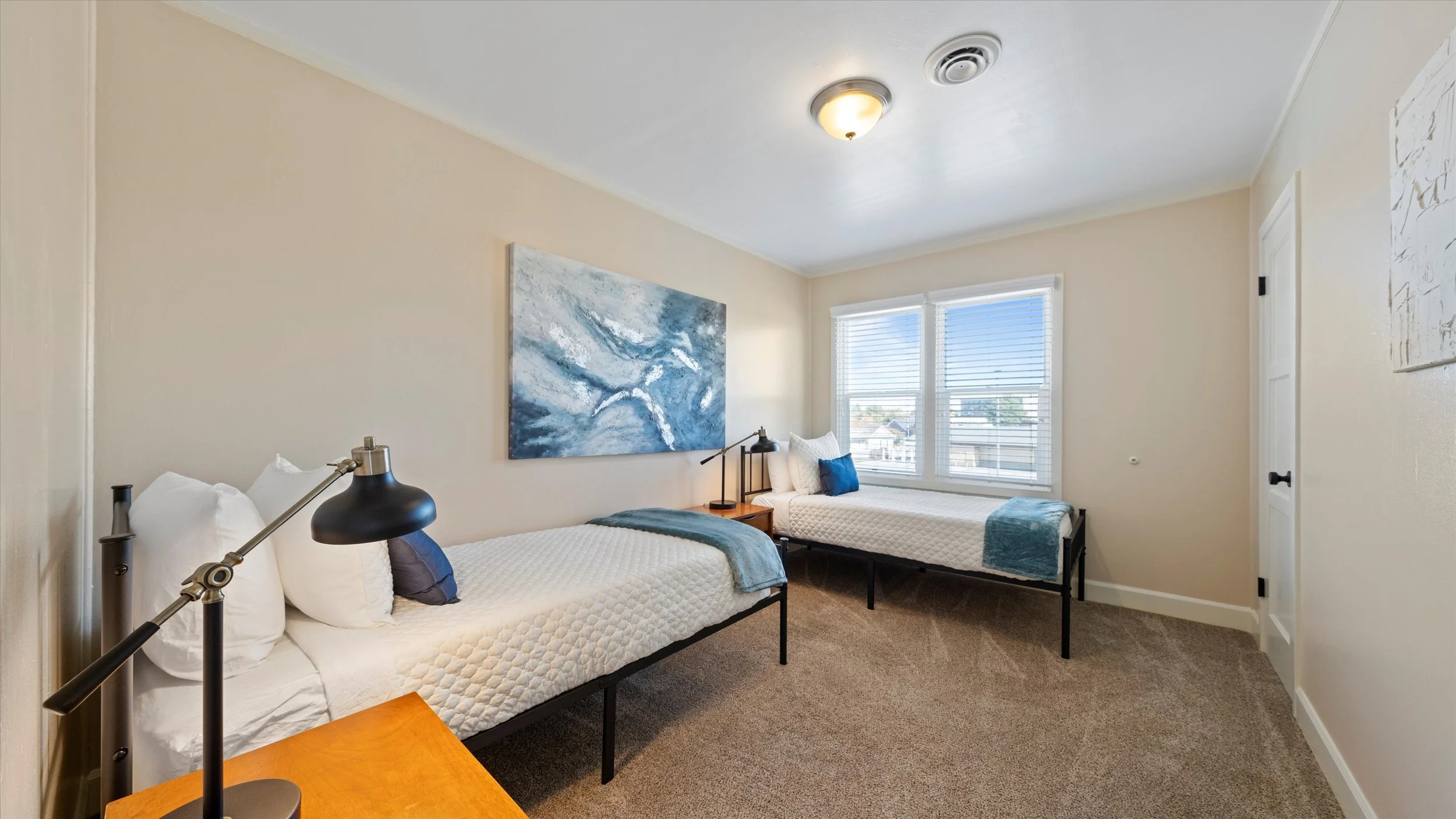 A bedroom with two twin beds, each with white quilts and pillows, a wooden nightstand between them with a black desk lamp, and a large window with blinds allowing natural light. The walls are painted light, and there's abstract blue artwork hanging a