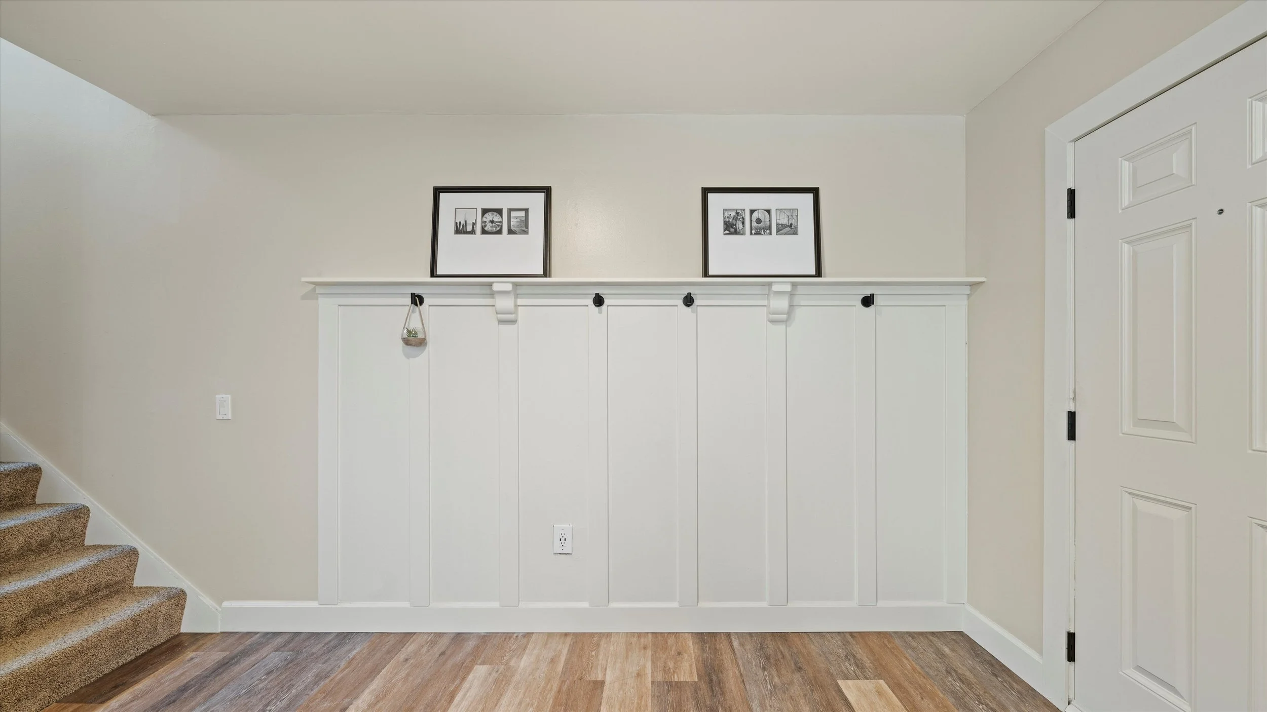 Interior of a home with a white wall, two black-framed photos, a shelf with hooks, and a white door to the right.