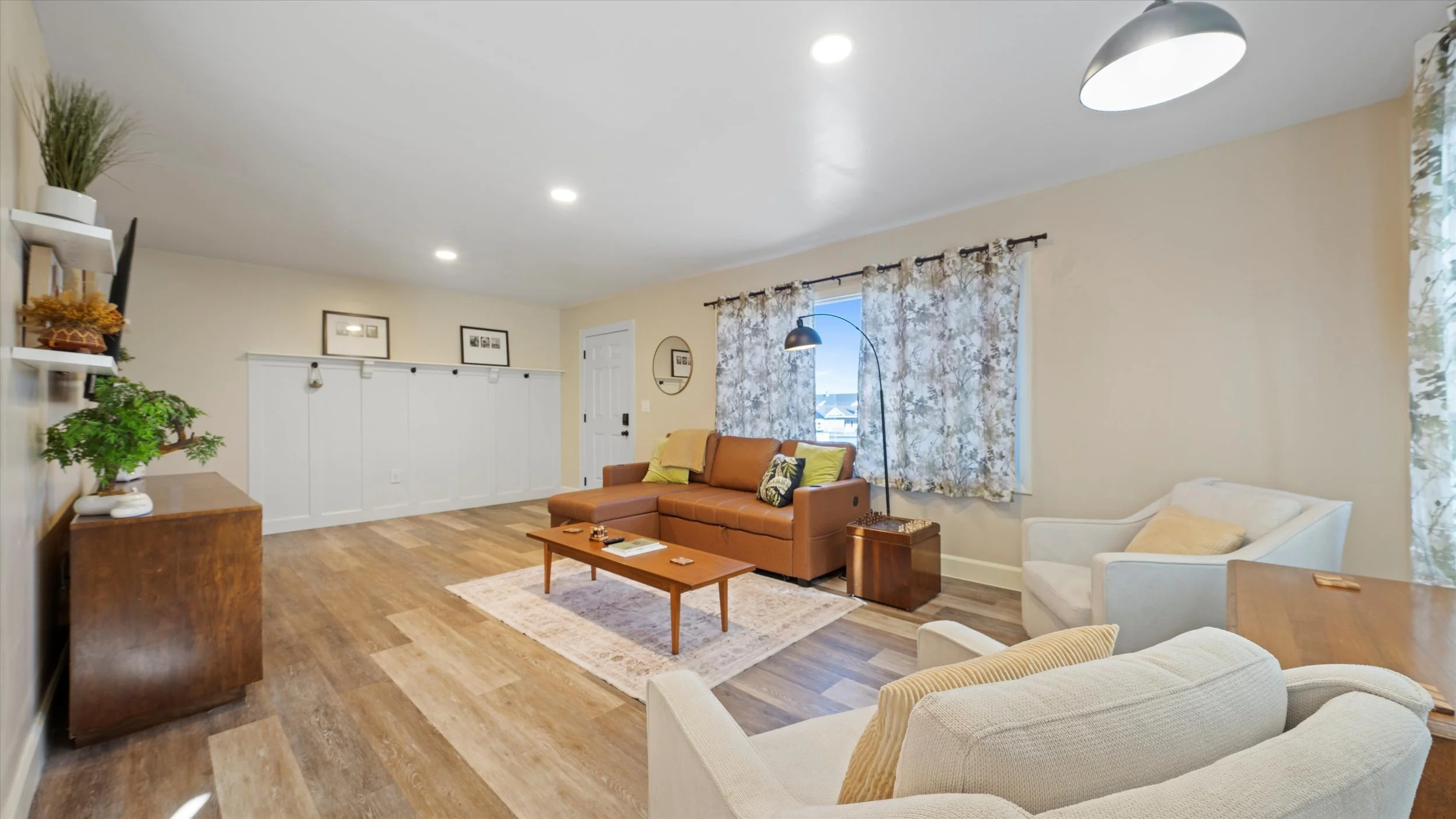 A living room with hardwood flooring, beige walls, and multiple windows with floral curtains. It features a brown leather sofa, a cream armchair, a white armchair, a wooden coffee table, a standing floor lamp, and decorative plants and pictures.