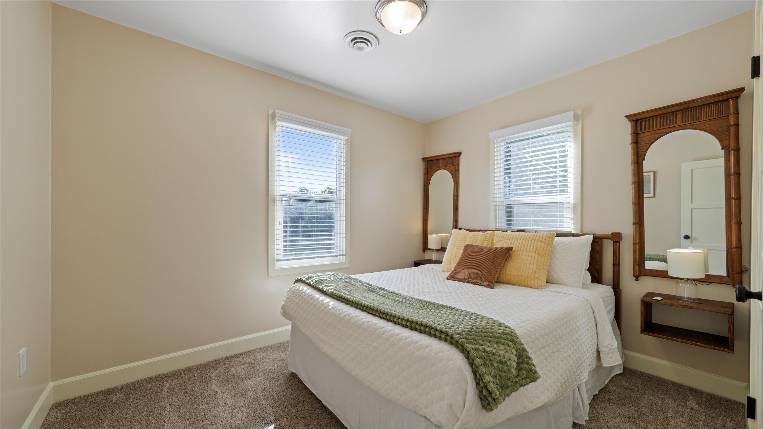 A bedroom with two windows, a bed with white linens and several pillows, and a wooden mirror and nightstand.