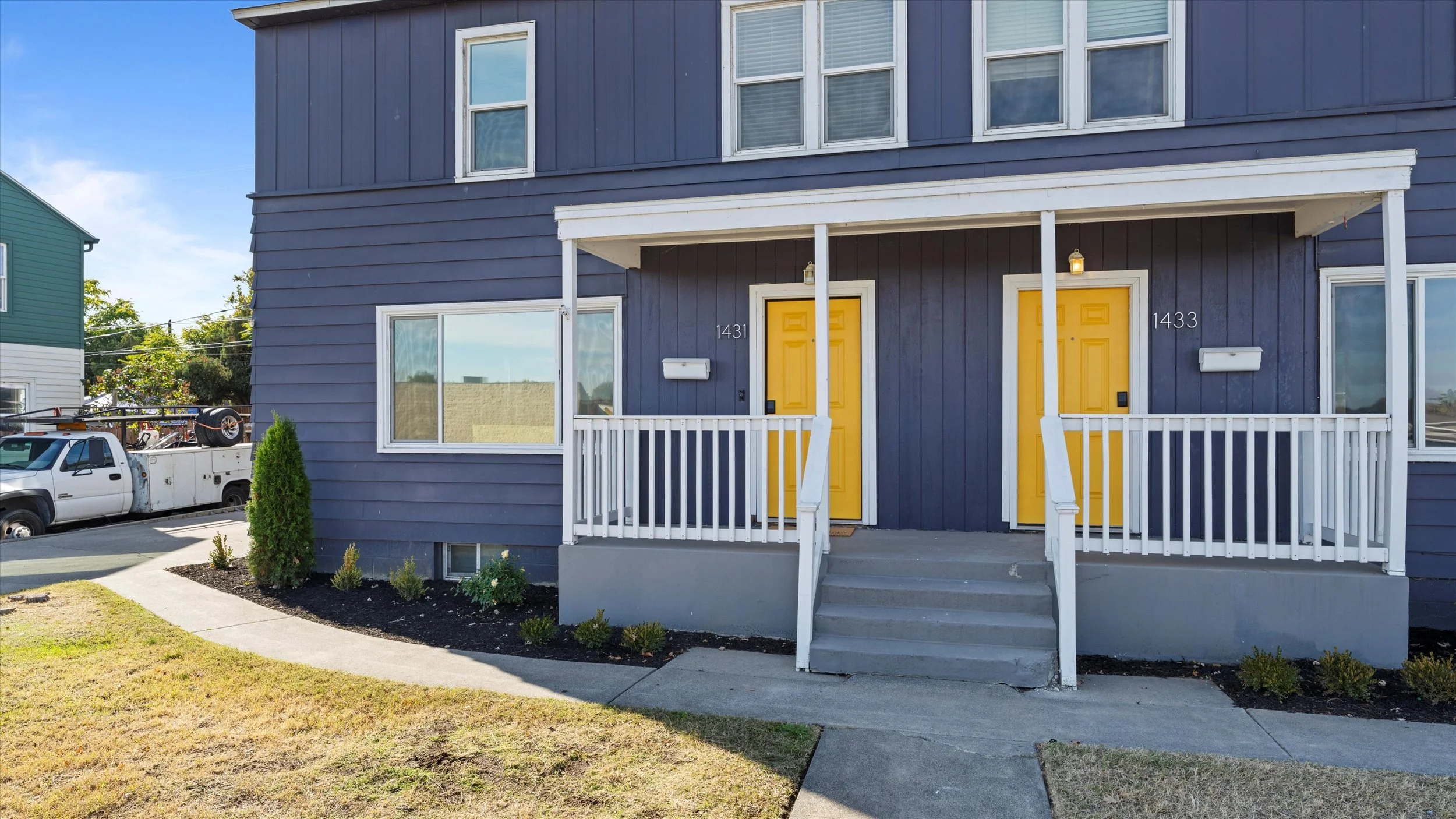 Front view of two blue townhouse units with yellow doors, white railing, and steps leading to the entrances. There is a small landscaped area with bushes and grass in front, and a sidewalk. A white truck is parked in the background.
