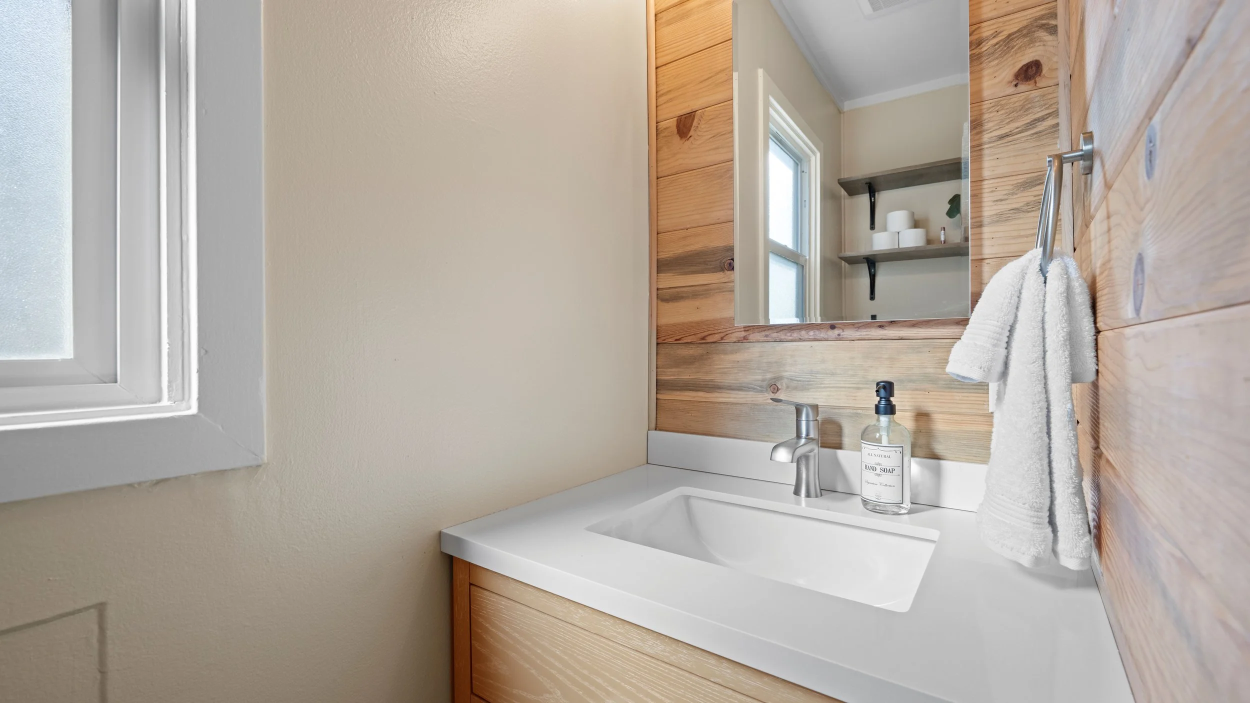 A bathroom sink area with a white countertop, a silver faucet, and a clear soap dispenser labeled 'HAND SOAP'. A towel is draped over the edge, and a mirror reflects shelves with toilet paper rolls. There is a window to the left, and the wall behind the sink is partially tiled with wood planks.