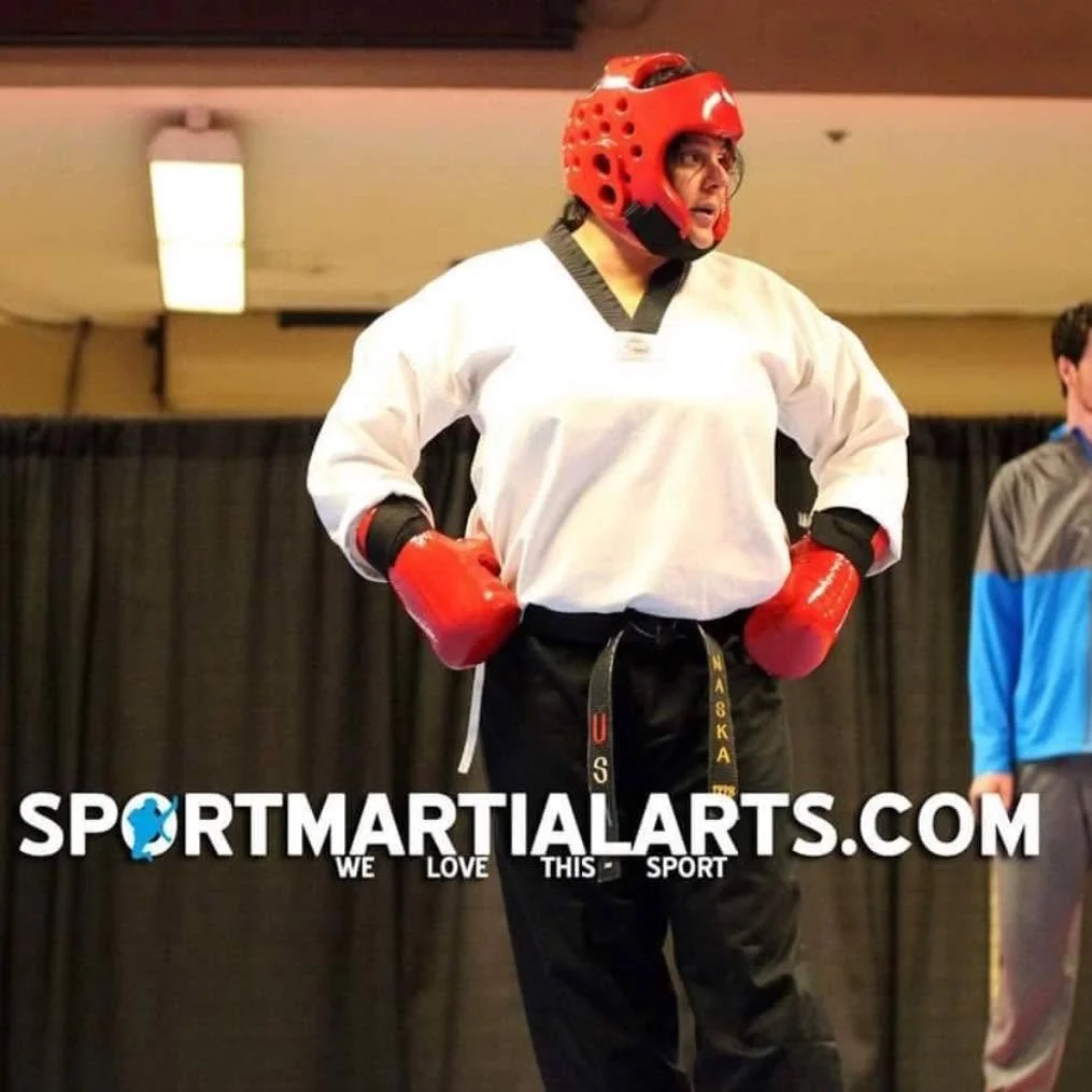 A woman in martial arts gear, including a red helmet, red gloves, a white martial arts uniform, and black martial arts pants, standing with hands on hips in an indoor setting.