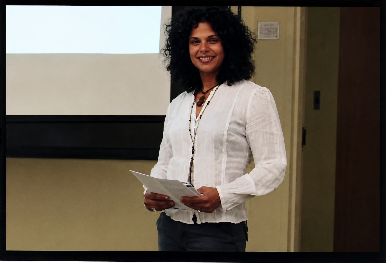 A woman with curly black hair smiling while holding a piece of paper, standing in front of a white projection screen in a conference or classroom setting.