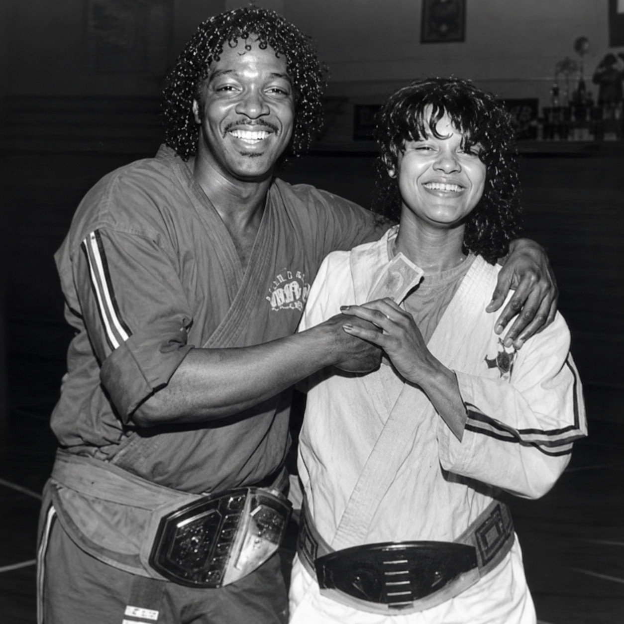 Two martial artists, a man and a woman, smiling and shaking hands, both wearing championship belts, in a gym or dojo setting.