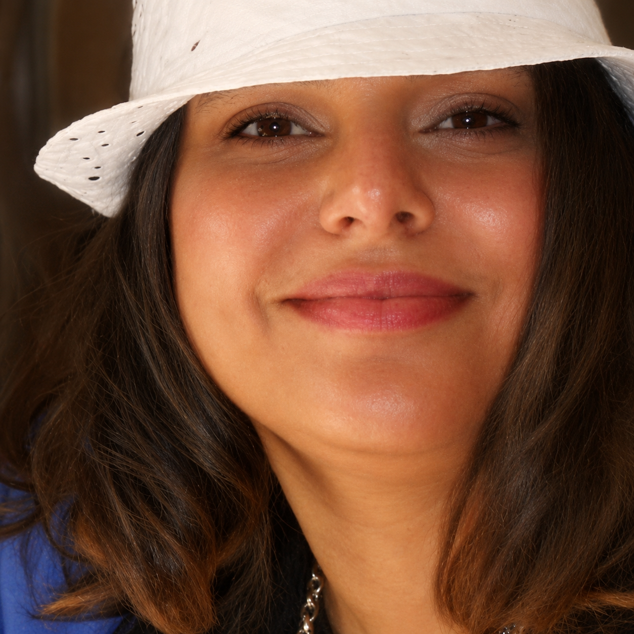 Close-up of a woman smiling, wearing a white straw hat with small holes, dark brown wavy hair, brown eyes, and minimal makeup.