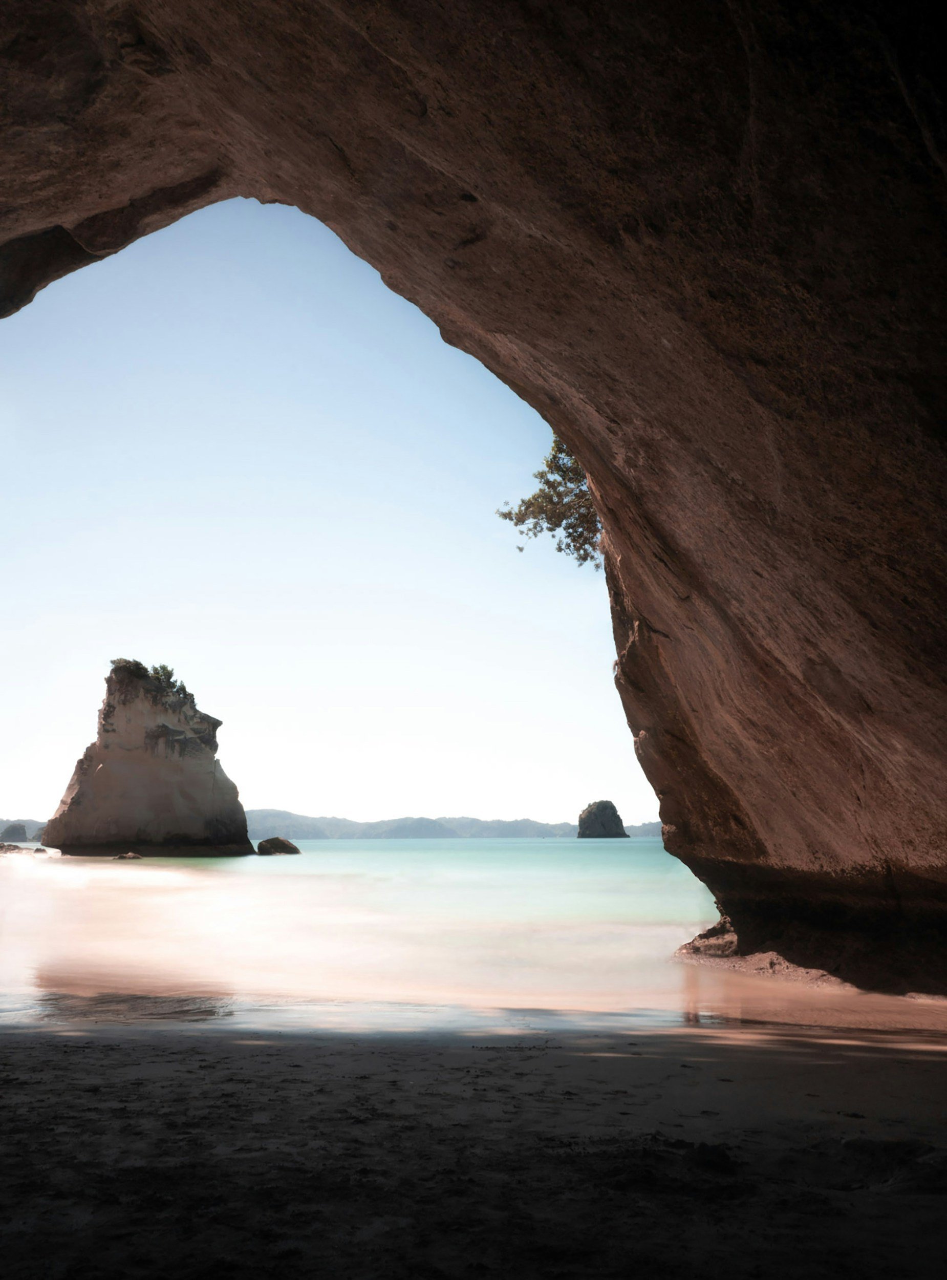 View of a beach with cliffs and rock formations seen through a large cave opening at sunset or sunrise.