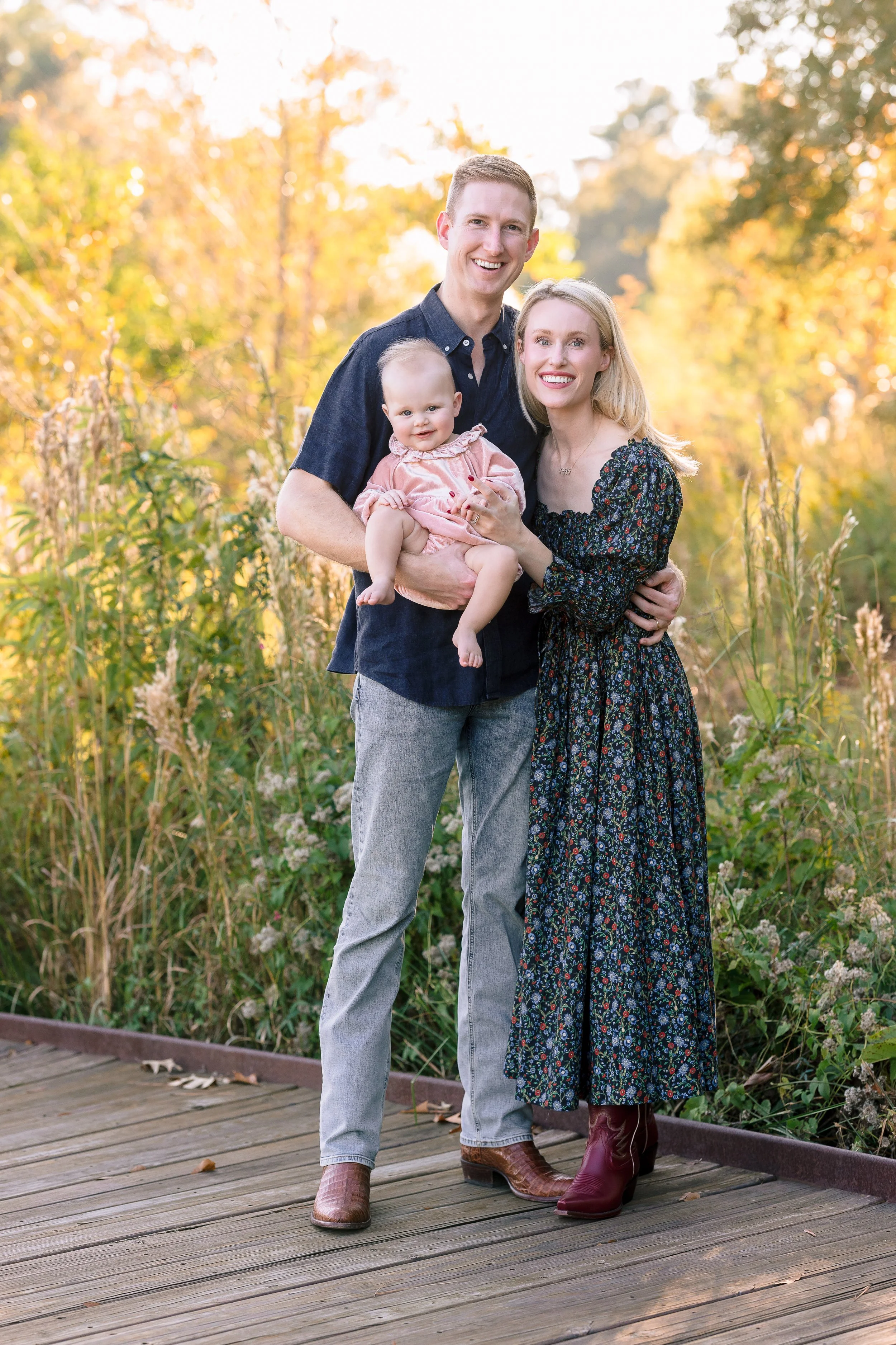 A family of three standing on a wooden pathway outdoors during autumn, with trees and golden foliage in the background. The man is wearing a dark blue shirt and gray jeans, holding a young child in pink clothing, and the woman is wearing a black floral dress with purple boots, smiling at the camera.