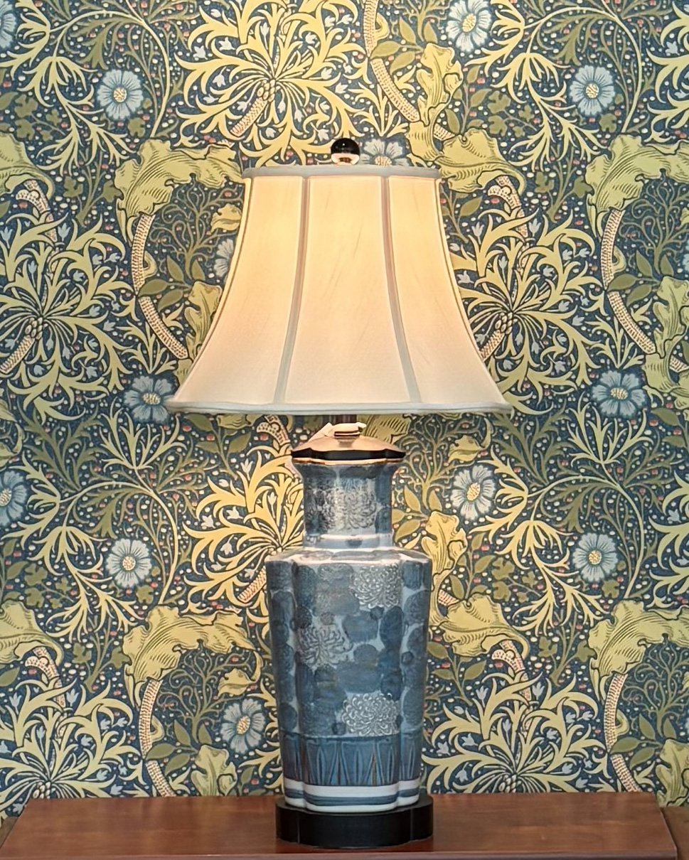 A decorative blue and white ceramic table lamp with a cream-colored lampshade on a wooden surface, against a navy blue wallpaper featuring yellow and blue floral and foliage patterns. Evelyn's Emporium in Saint Paul, MN.
