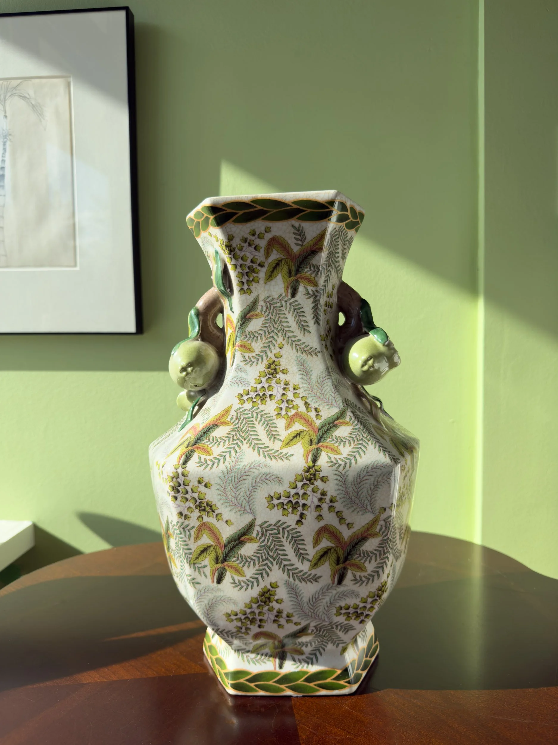 Chinese-inspired porcelain hexagonal vase with fern design and pomegranate handles, Evelyn's Emporium, Saint Paul, MN. The vase has a leaf and vine pattern, and is resting on a wooden table with a green wall background.