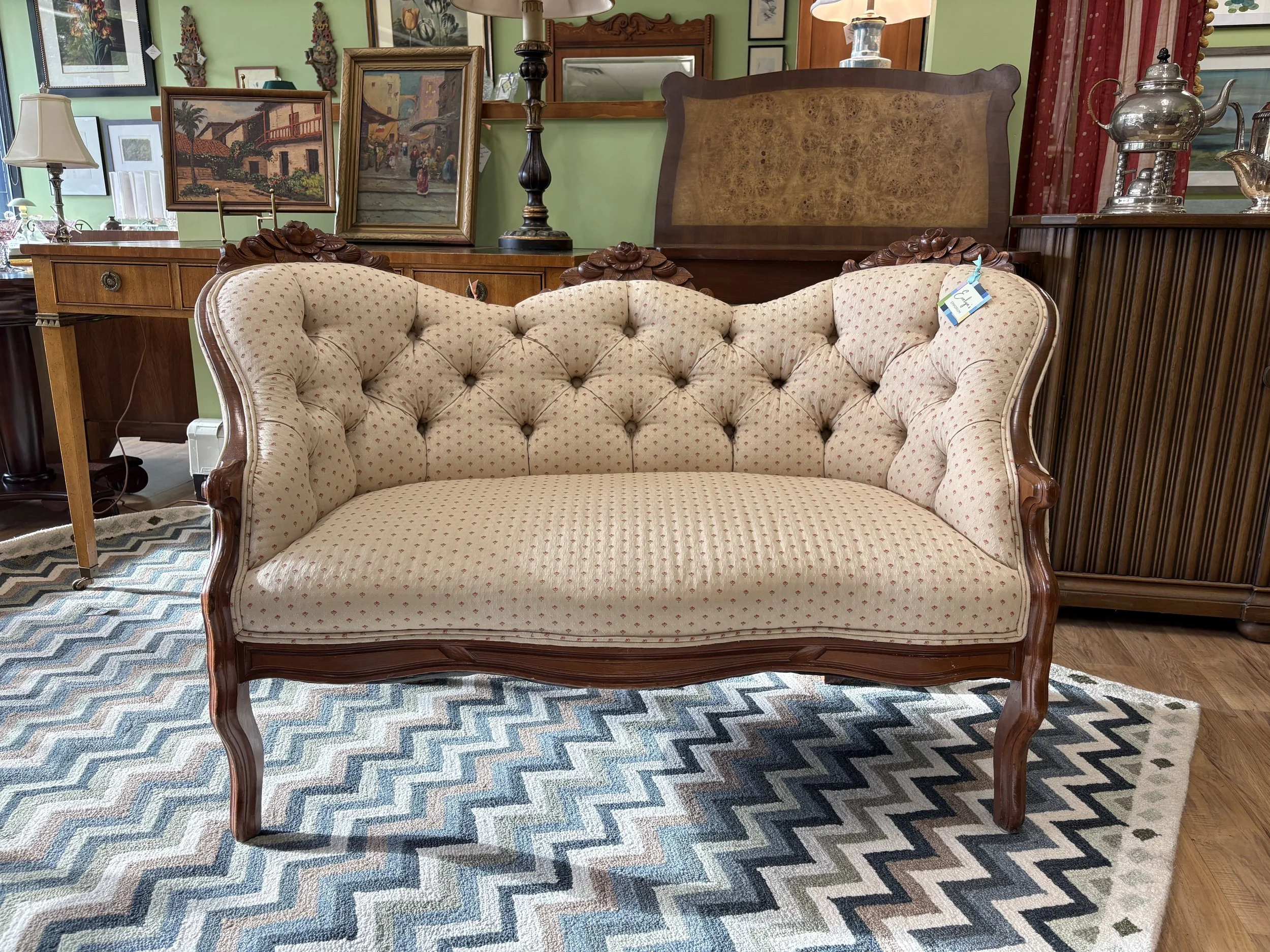 A vintage-style cream-colored sofa with tufted upholstery and wooden carved armrests and legs, positioned on a geometric-patterned rug. Displayed in Evelyn's Emporium with framed paintings, lamps, and decorative items in the background.