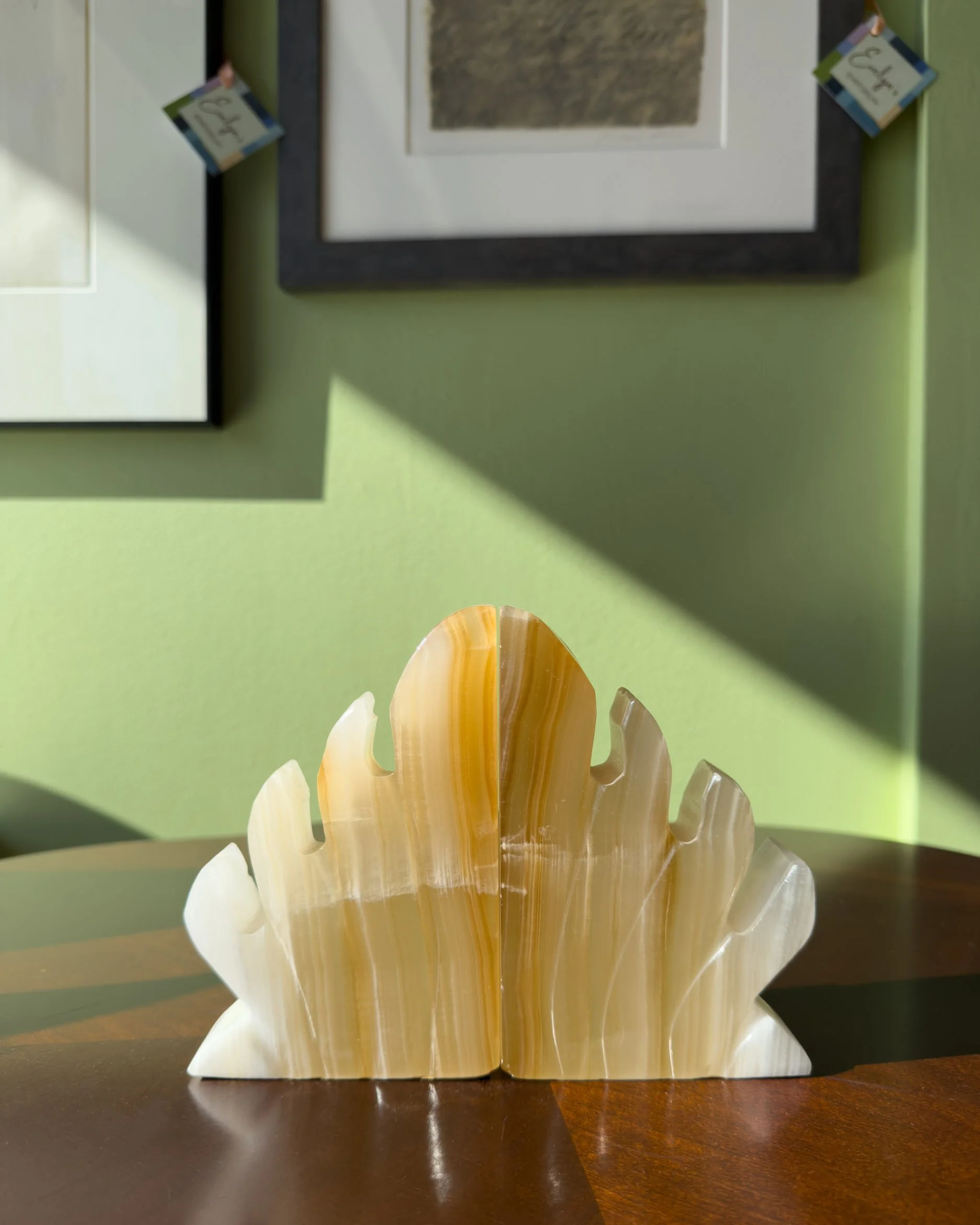 A decorative set of polished onyx bookends with swirling patterns in shades of amber, cream, and white, resembling flower petals, is displayed on a dark wooden table against a green wall with framed artwork. Evelun's EMporium in Saint Paul, MN.