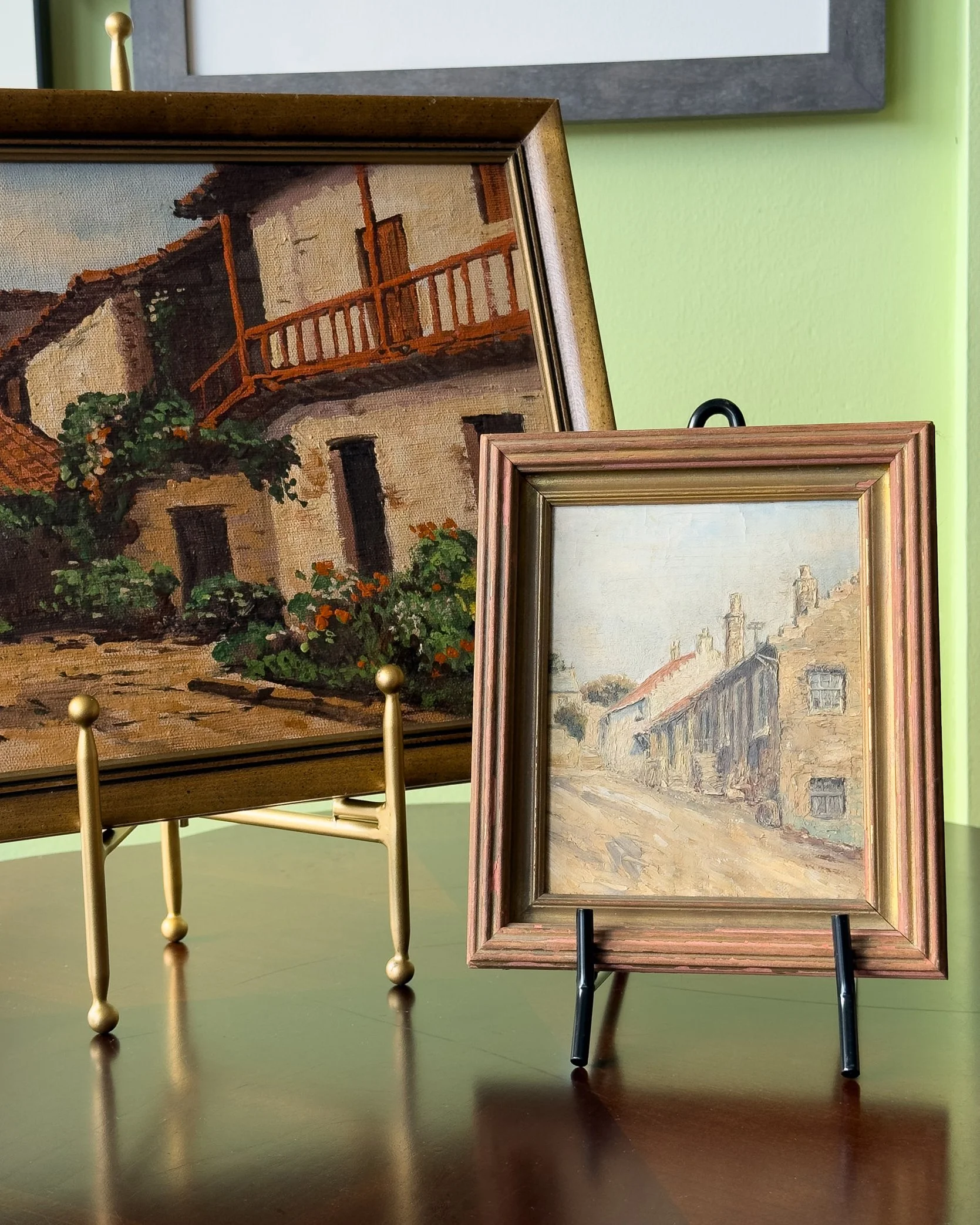 Two framed paintings are set on small easels on a green table. The paintings depict outdoor scenes, with one showing a European street and the other a countryside village.