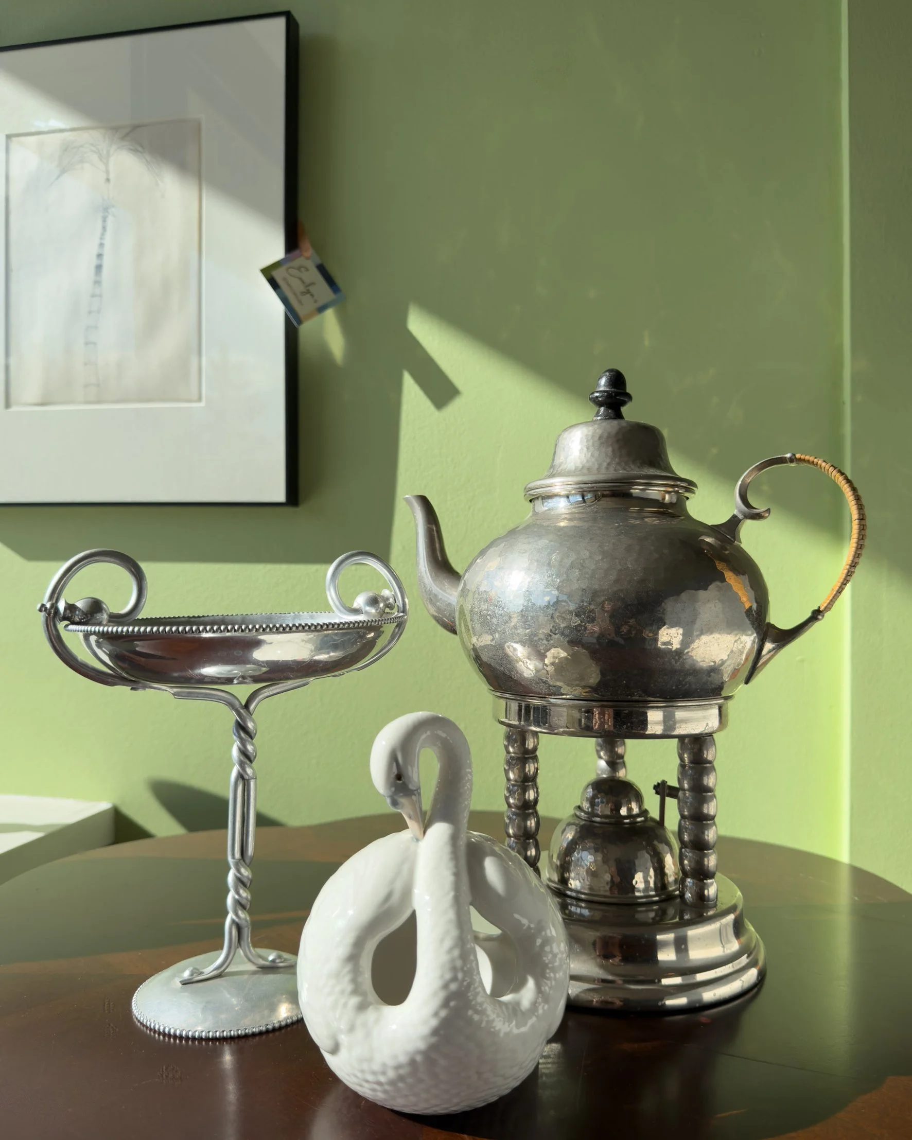 Three decorative items on a dark wooden table against a light green wall. Includes a white ceramic swan-shaped vase in the foreground, a vintage silver teapot with a woven handle, and a two-tier silver candy tray. Evelyn's Emporium in Saint Paul, MN.