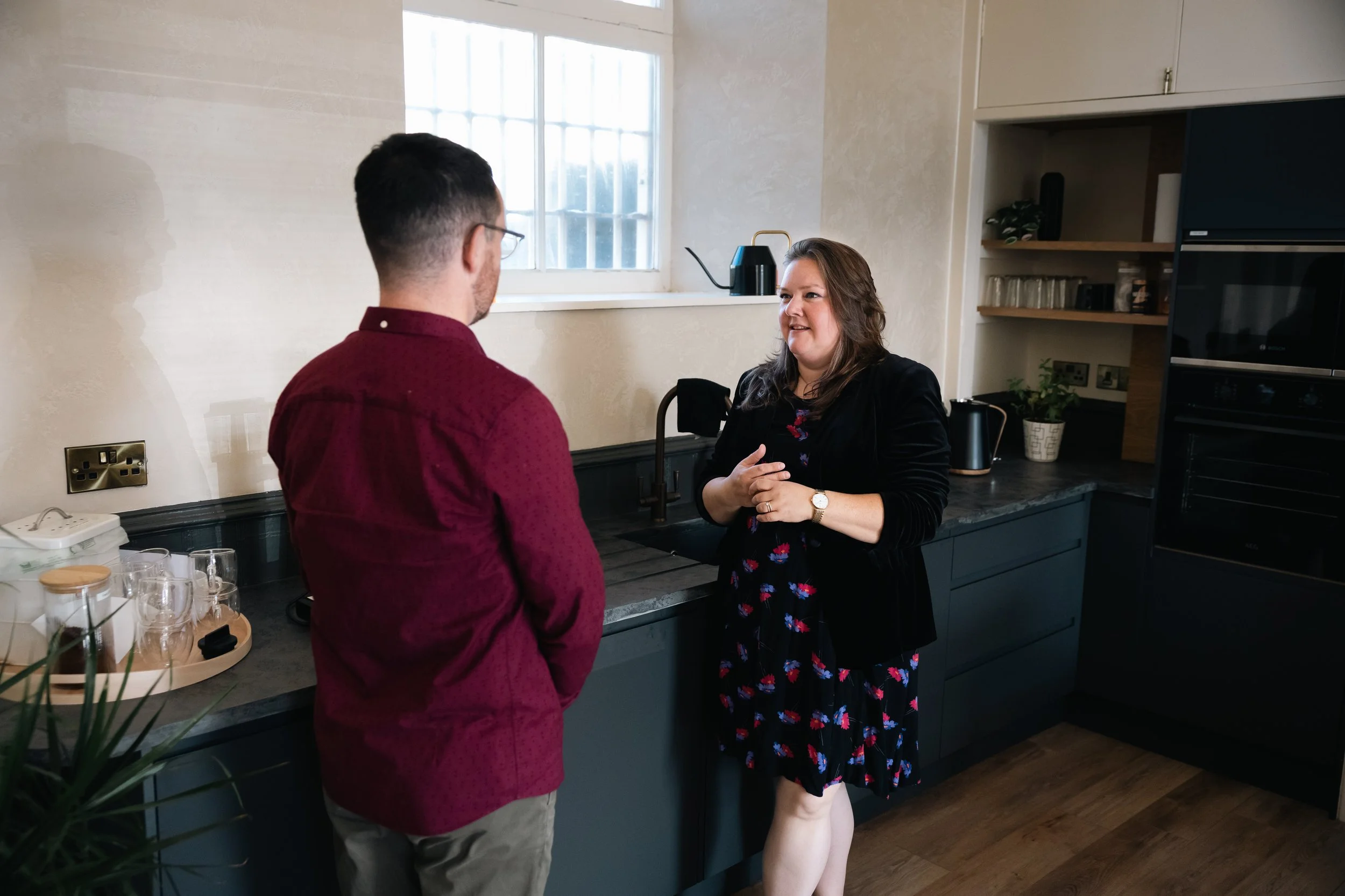 A man and a woman are having a conversation in a kitchen. The man is wearing a maroon shirt and glasses, with a short haircut. The woman has long brown hair and is wearing a black jacket over a floral dress. The kitchen has blue cabinets, a window, and various kitchen items on the counter.