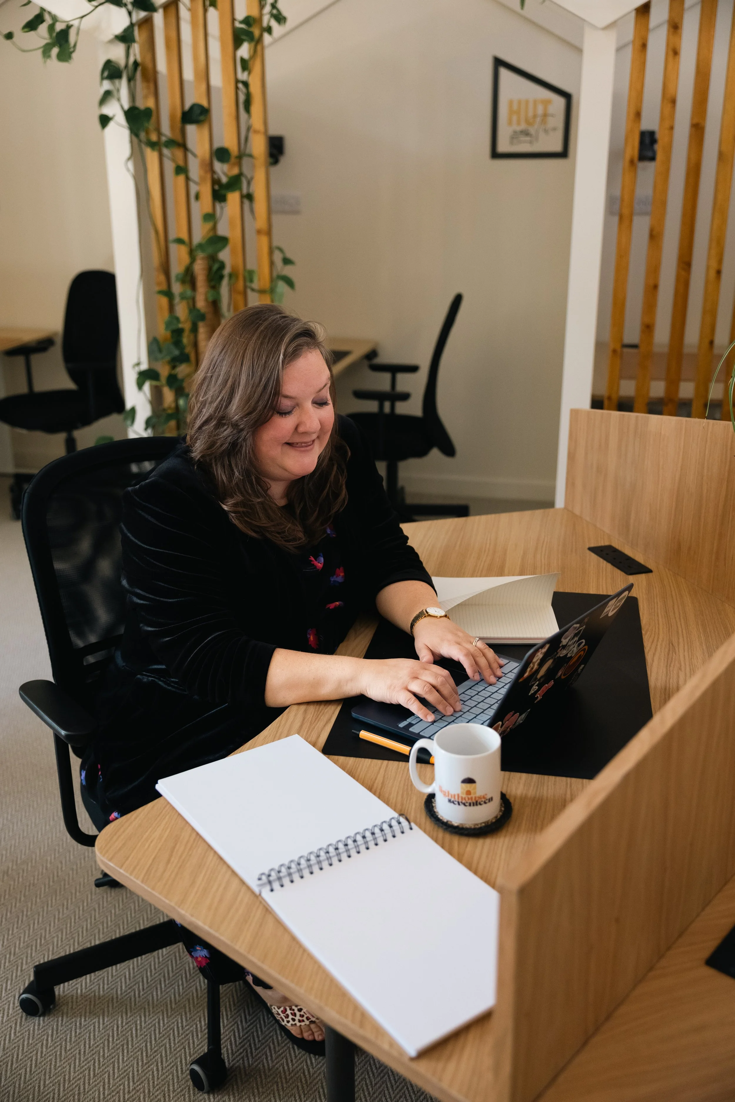 A woman with shoulder-length brown hair sitting at a wooden desk, typing on a laptop. The desk has a white notebook, a white coffee mug, and an orange pen. The background features black office chairs, wooden slats, potted plants, and framed wall art.