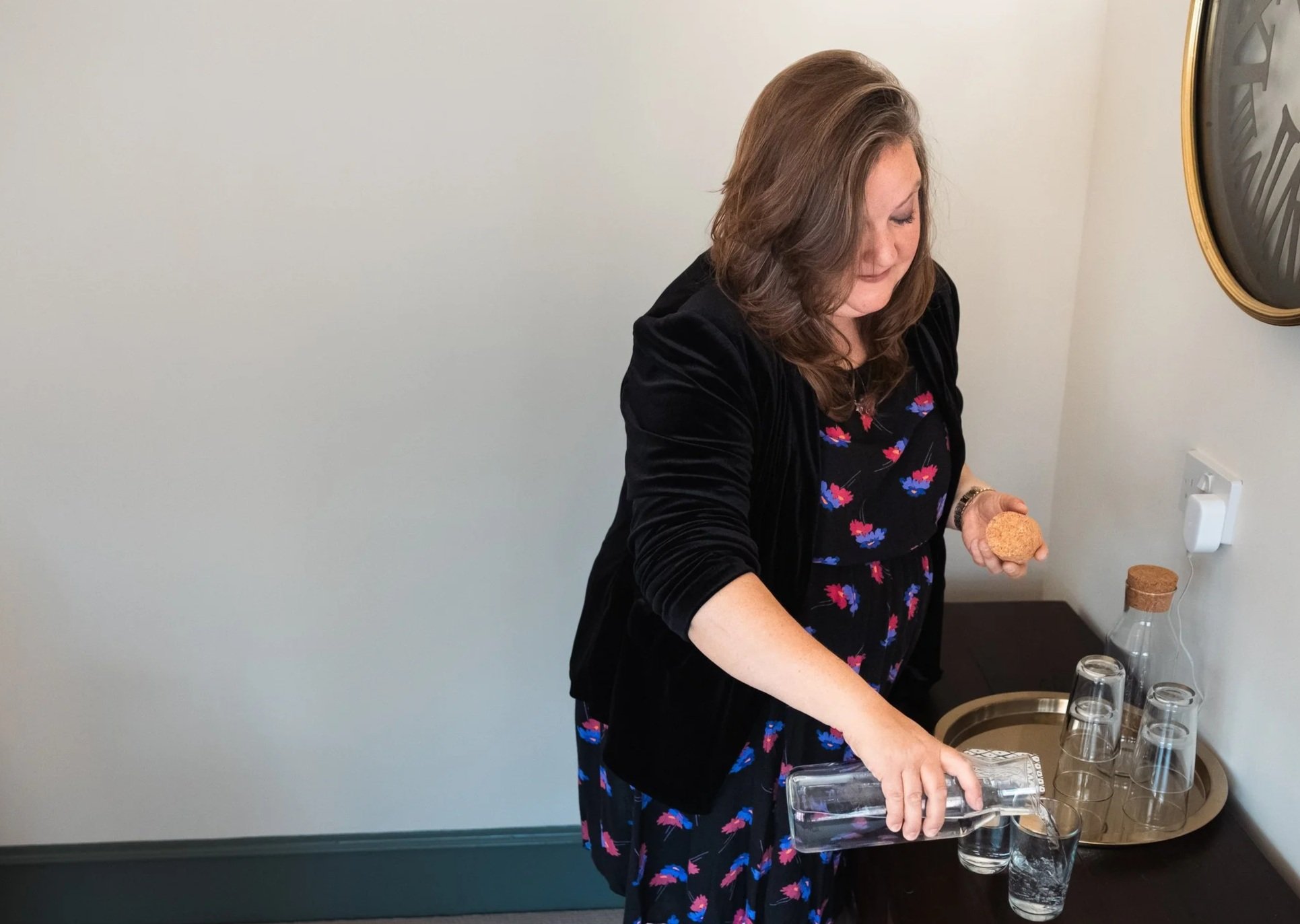 A woman with shoulder-length brown hair wearing a black cardigan and a dress with a floral pattern pouring a drink into clear glass cups on a table.