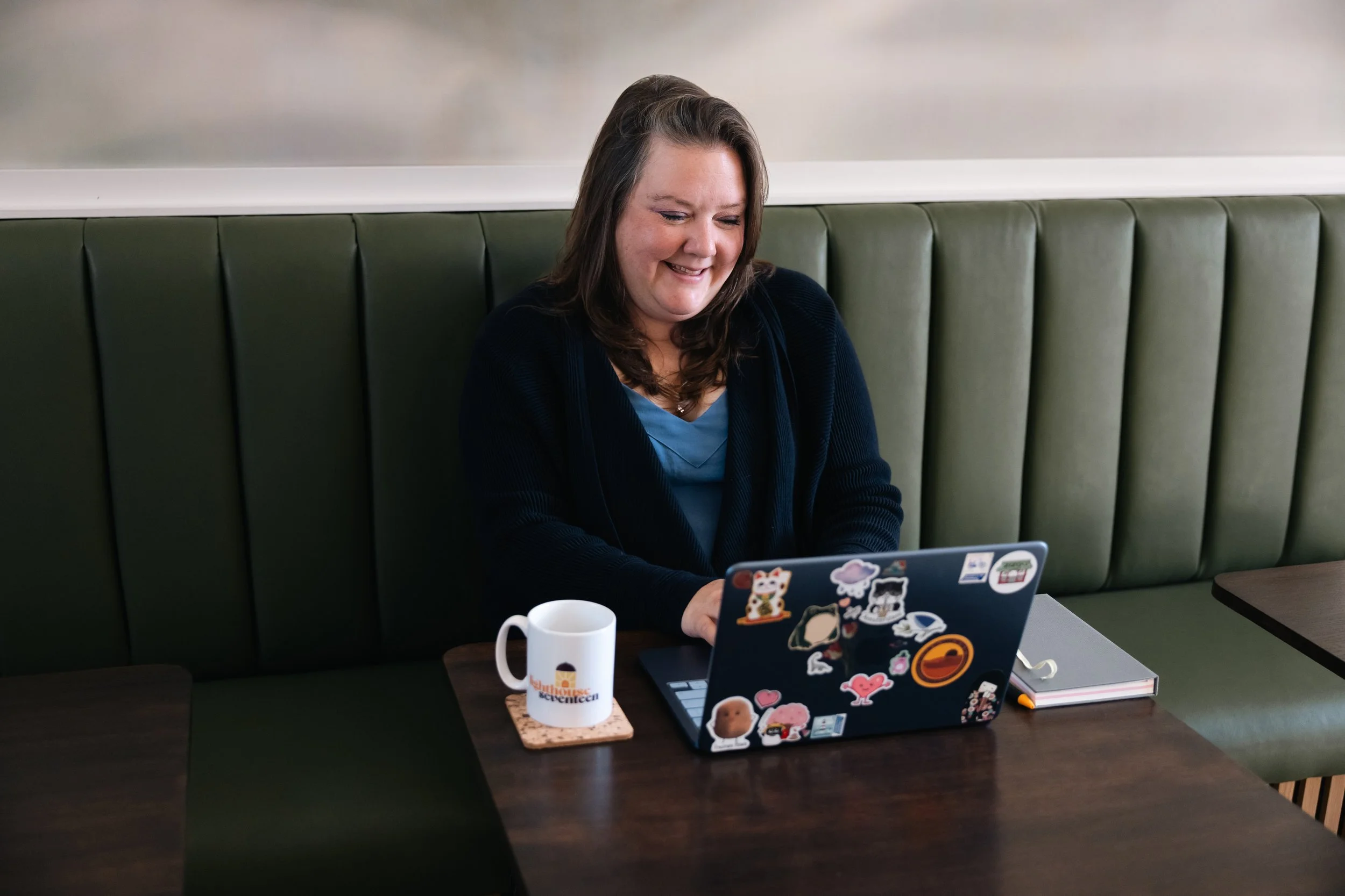 A woman with long brown hair, smiling while looking at her laptop covered in various stickers, sitting at a dark wooden table in a booth with green padded seating, a white mug, and a notepad nearby in a casual setting.