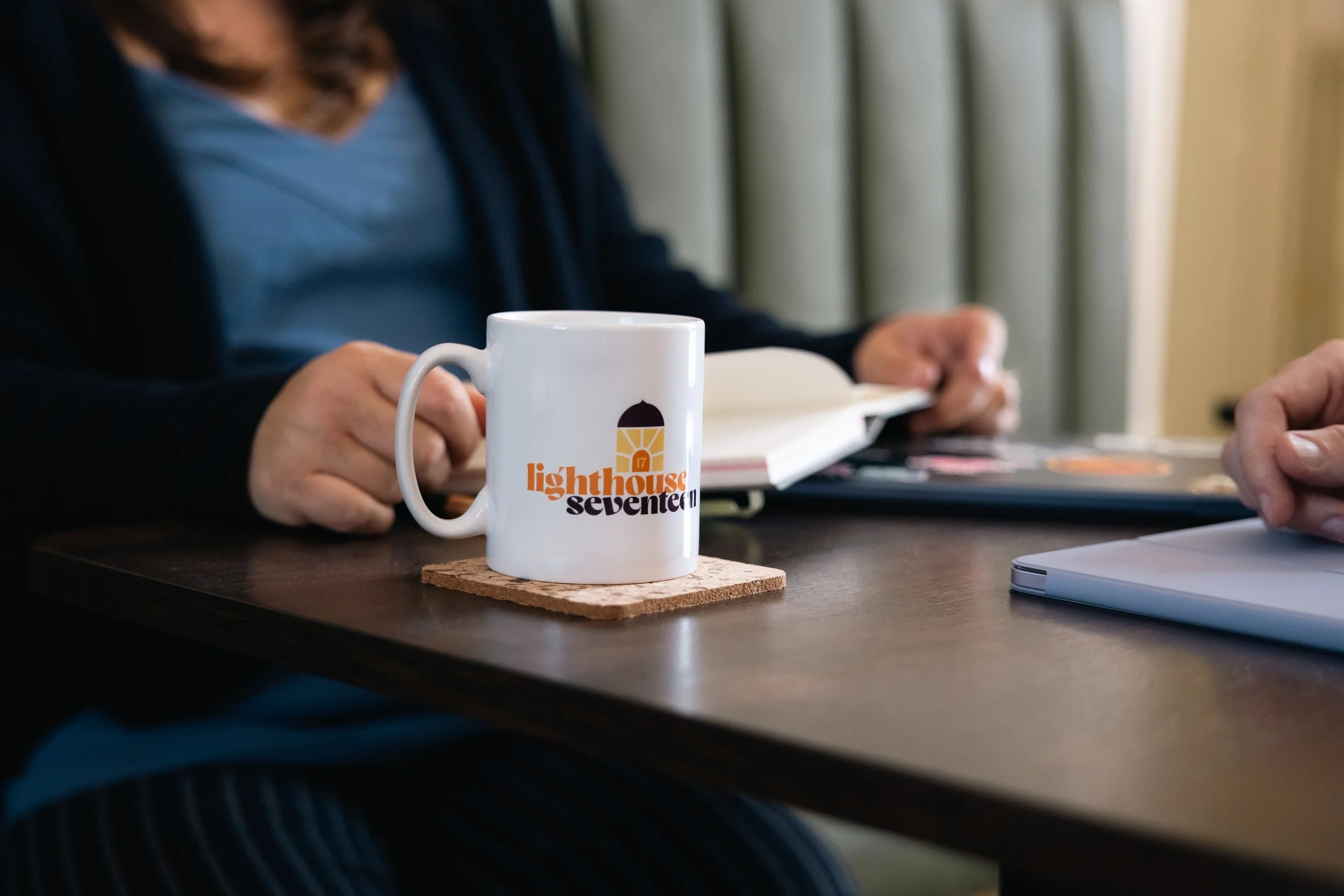 Close-up of a white coffee mug with the Lighthouse Seventeen logo, sitting on a cork coaster on a dark wooden table, with a person reading a book or magazine in the background.
