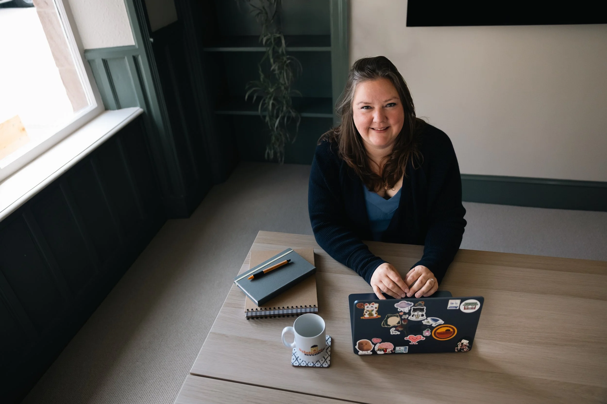 A woman sitting at a light-colored wooden desk, smiling at the camera, with a black notebook, pen, and a silver laptop decorated with stickers, a coffee mug, and a mobile phone on the desk. There is a window to her left and a dark-colored cabinet with a plant behind her.