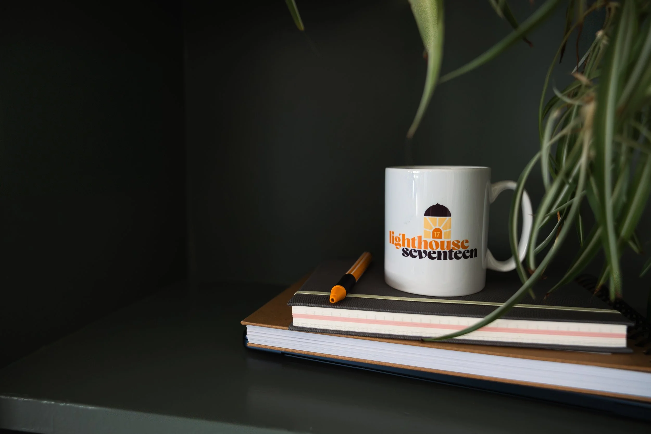 A white mug with a logo and text, resting on a stack of notebooks and a black pen, next to a green plant on a dark surface.