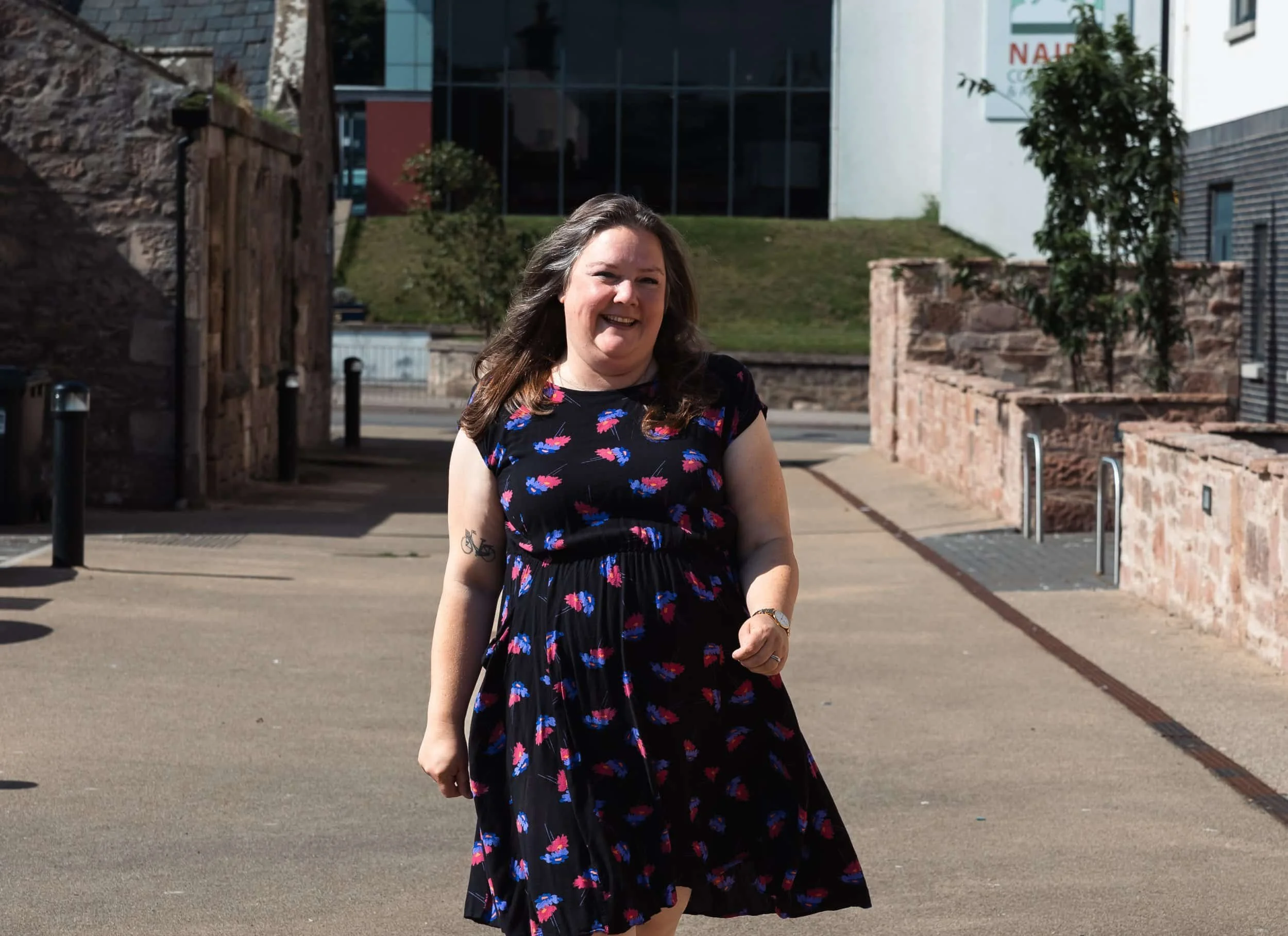 A woman with long brown hair, wearing a black dress with colorful floral patterns, is smiling while walking outdoors on a sunny day. In the background, there are brick walls, a grassy area, trees, and modern buildings.
