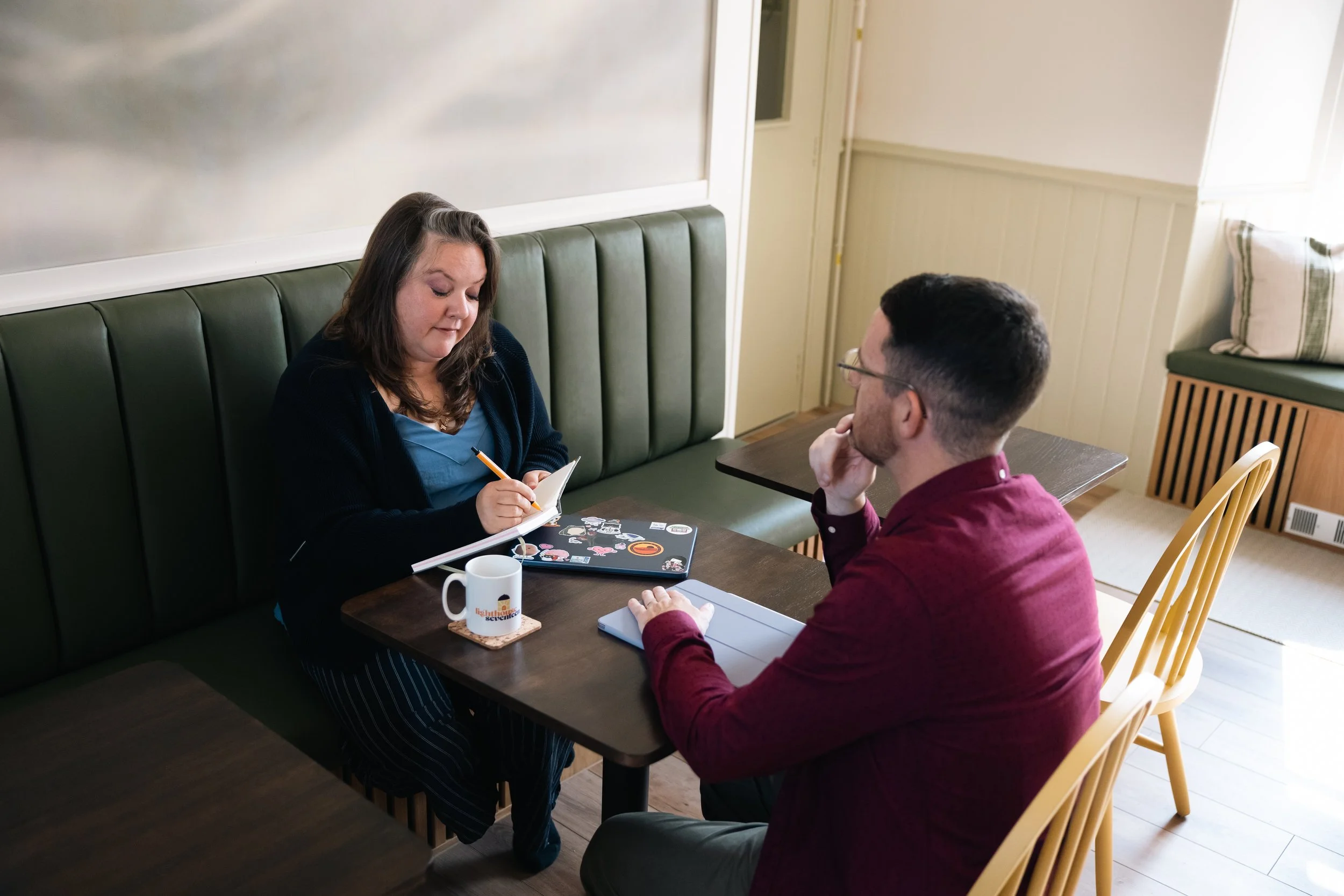 A woman taking notes across from a man with glasses in a cozy cafe or restaurant with wood panel walls, green cushioned seating, and a window with blinds.