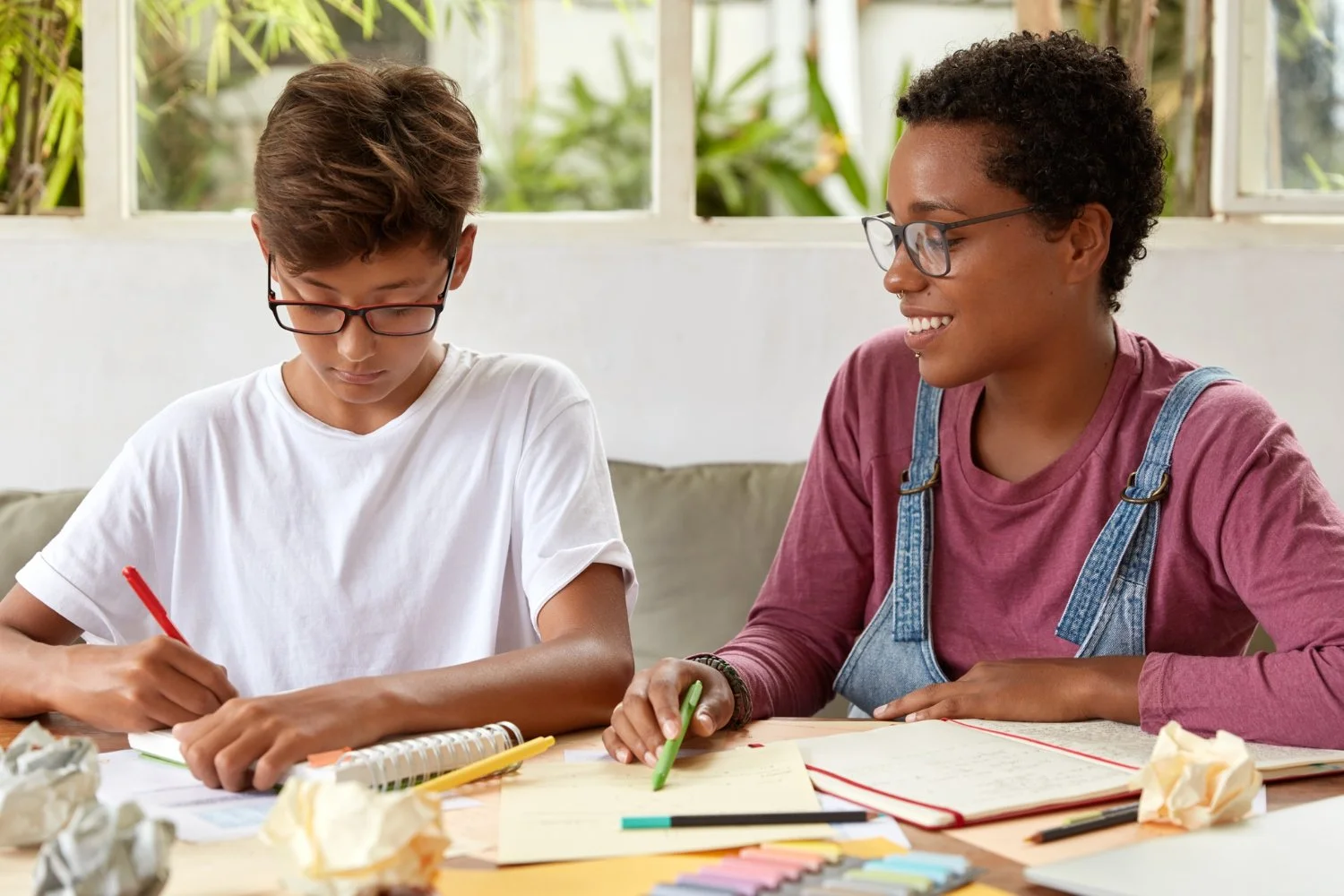 Two people, a young person and an adult, sitting at a desk with notebooks, papers, and crayons, engaging in an activity together in a bright room with large windows and green plants outside.