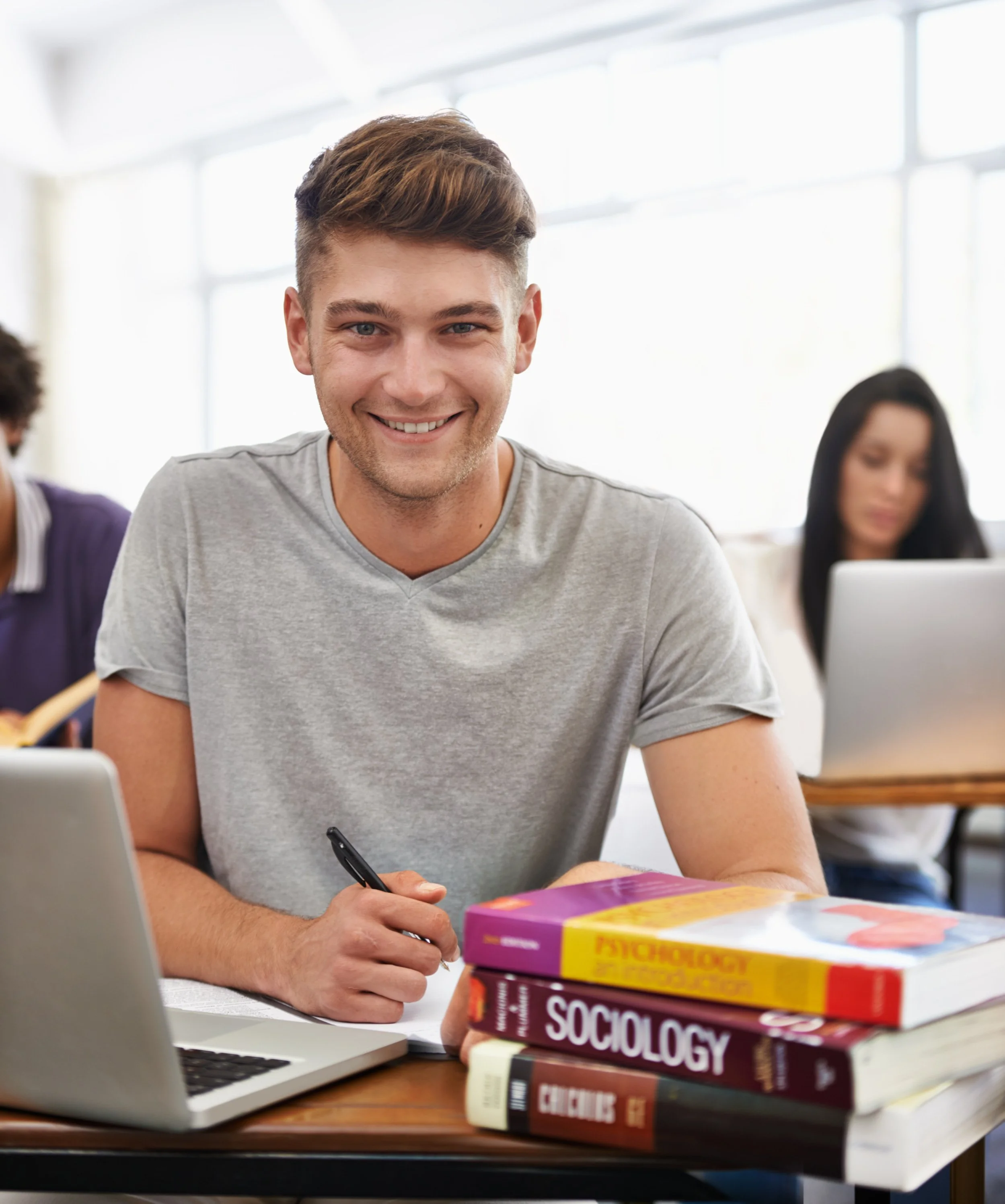 A young man with short brown hair and a gray t-shirt sitting at a desk with a smile, writing on a piece of paper with a pen. In front of him, there are several thick textbooks stacked, including titles like 'Sociology' and 'Psychology.' A laptop and a purple and yellow book are also on the desk, with a bright, window-lit classroom or study area in the background.