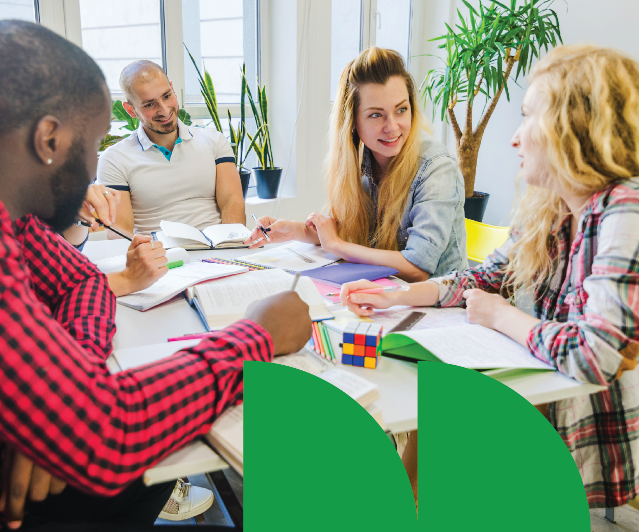 Group of young adults collaborating at a table in a bright, modern office, with books, notebooks, and a Rubik's cube.