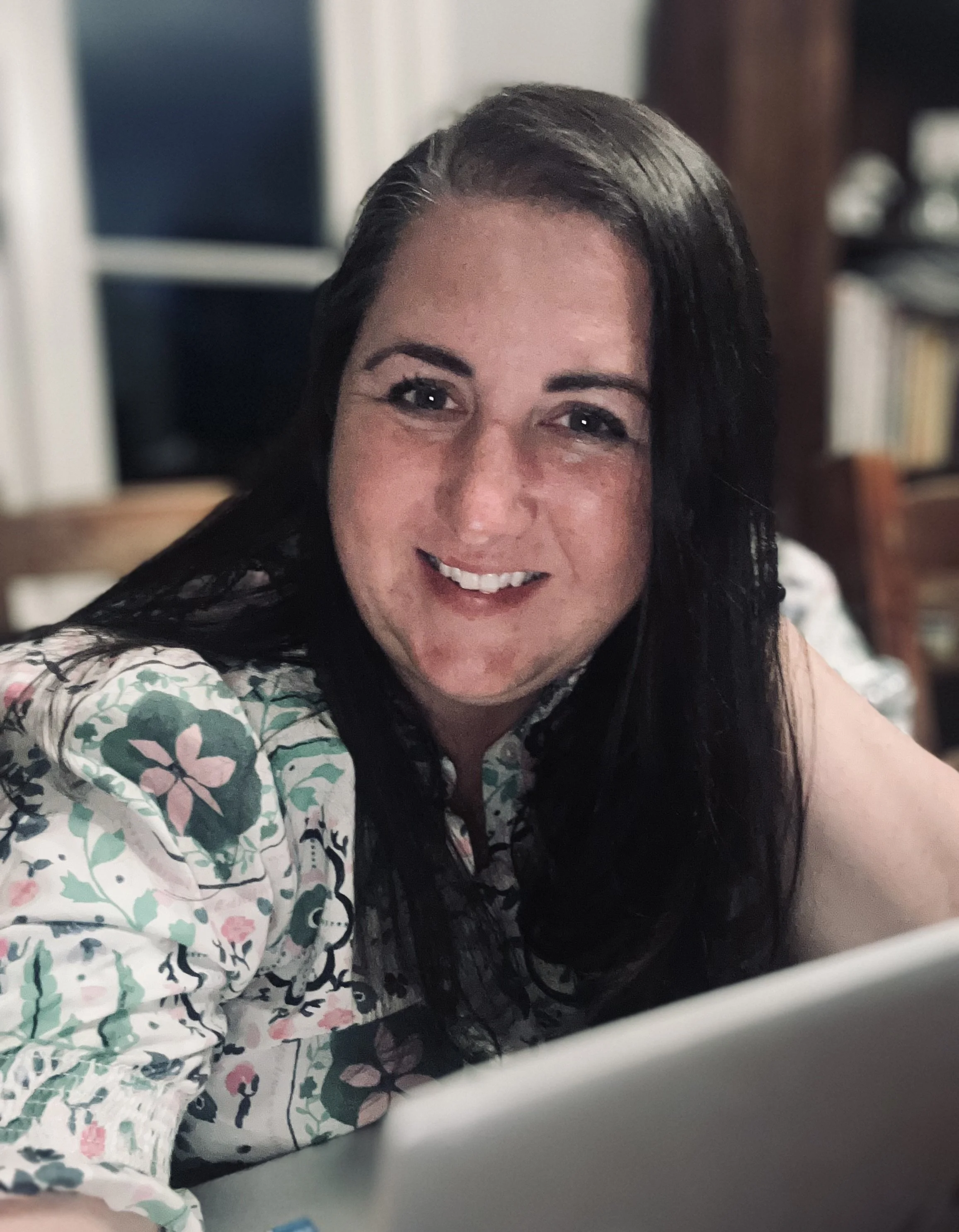 A woman with long dark hair smiling at the camera, wearing a white shirt with a colorful floral pattern, sitting behind a table.