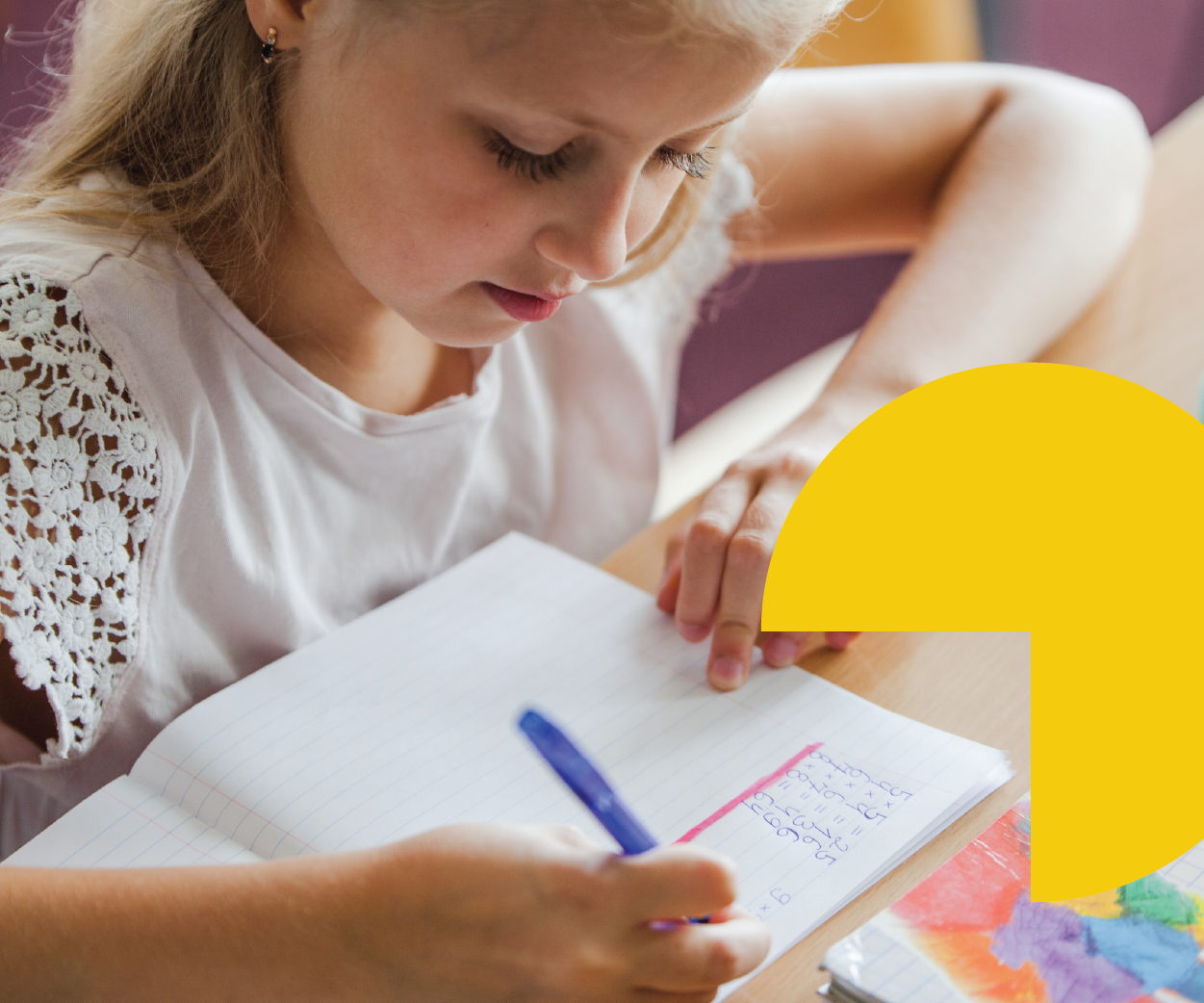 A young girl with blonde hair, wearing a white shirt with lace sleeves, is sitting at a wooden desk, focused on writing or solving math problems in a notebook with blue ink, a blue pen in hand, with colorful artwork nearby.