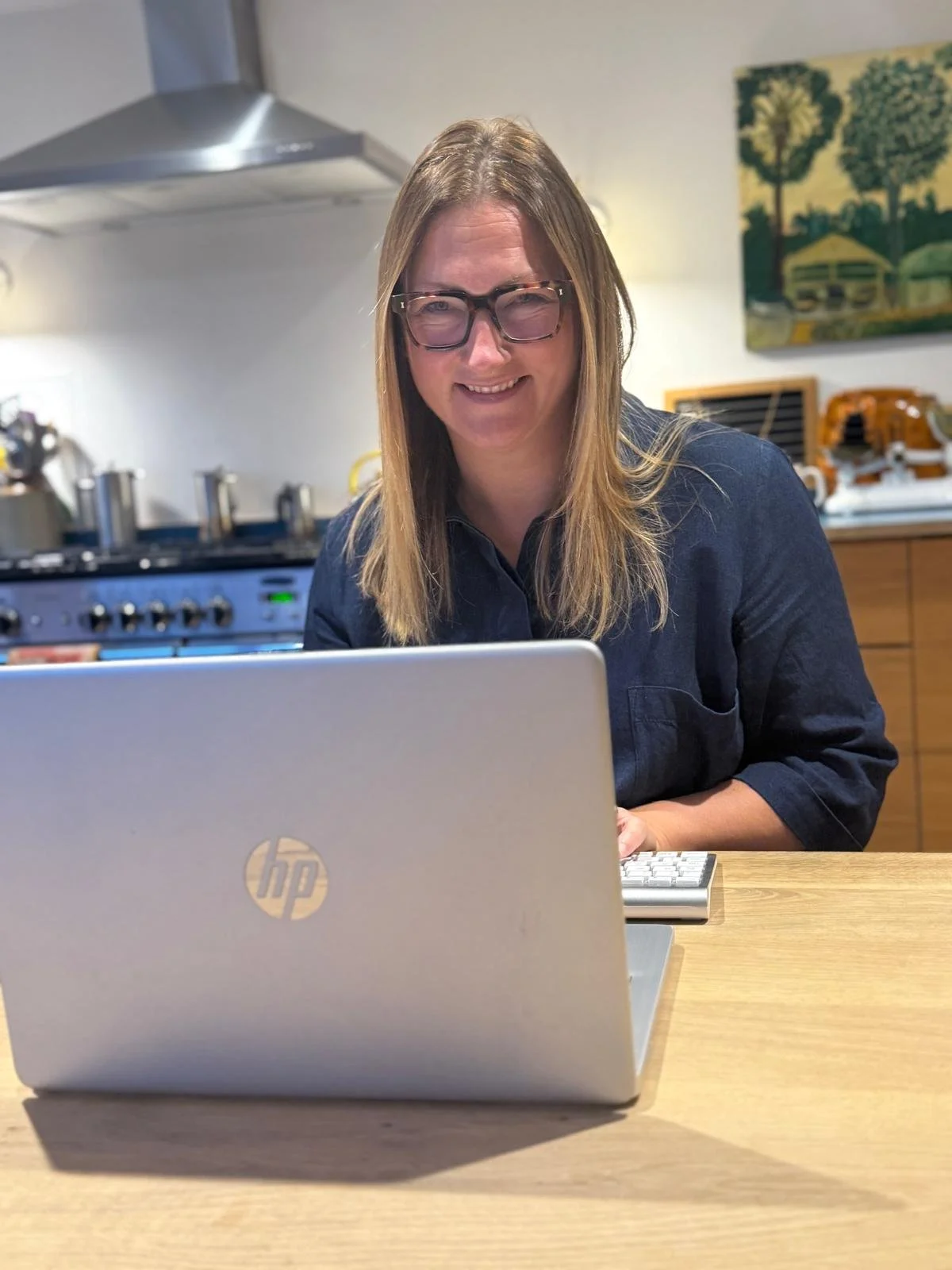 A woman with long hair and glasses smiling at a laptop in a kitchen.
