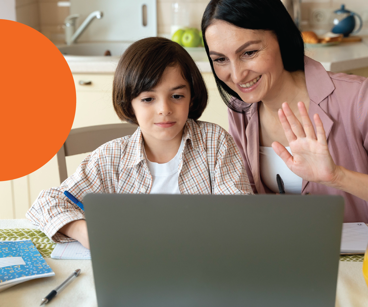 A woman and a young boy looking at a laptop screen together at a kitchen table, with the woman waving and smiling.
