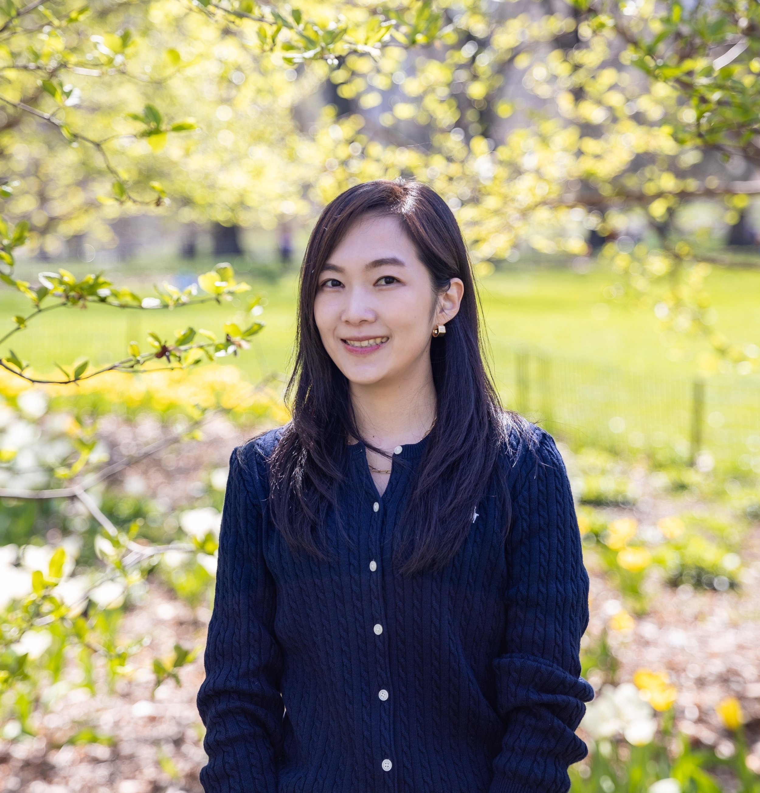 A woman with long dark hair and earrings standing outdoors in front of yellow-green foliage, wearing a navy blue knit sweater with white buttons, smiling at the camera.