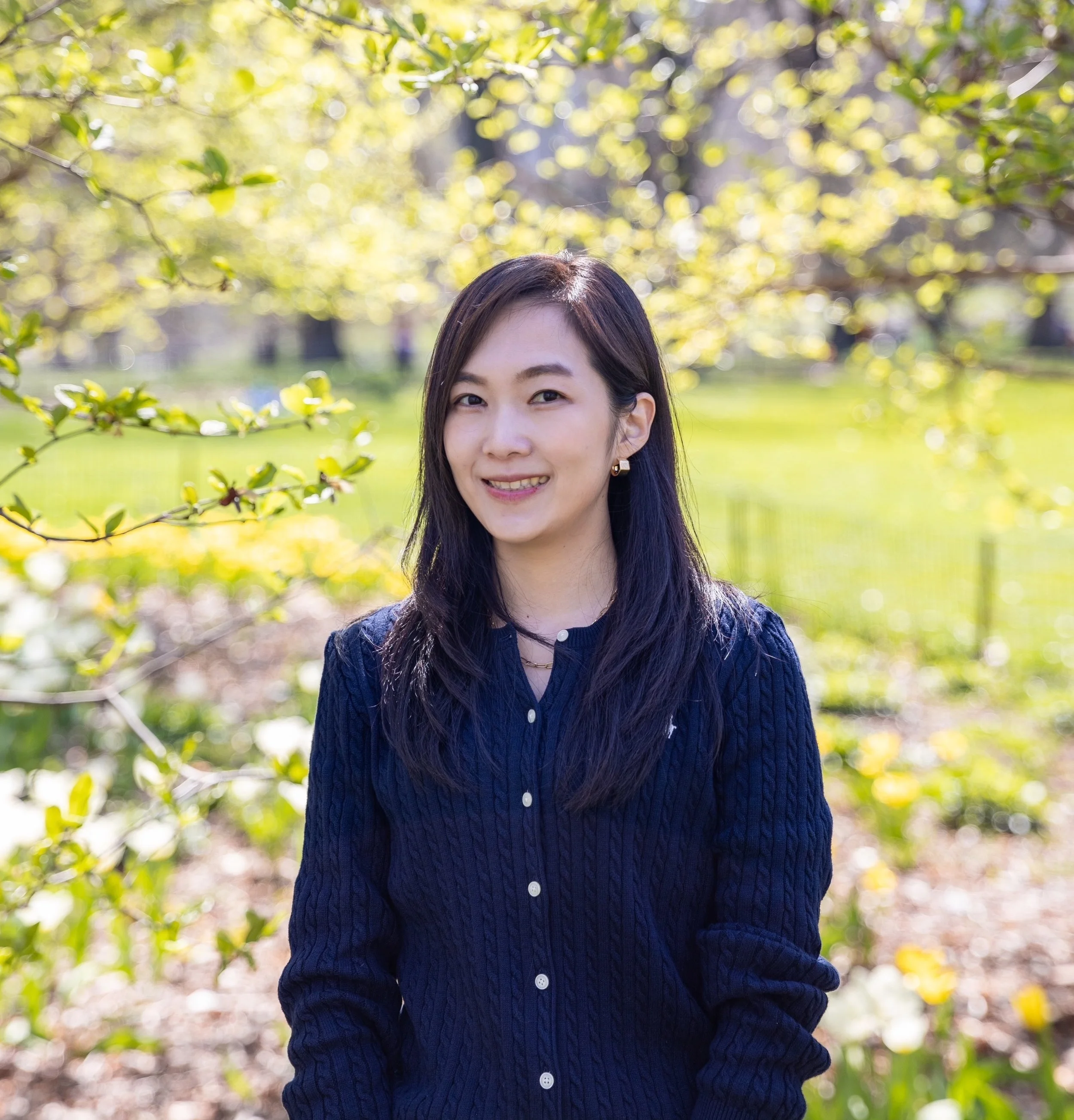 A woman with long dark hair and a denim shirt standing outdoors in front of green trees with sunlight filtering through